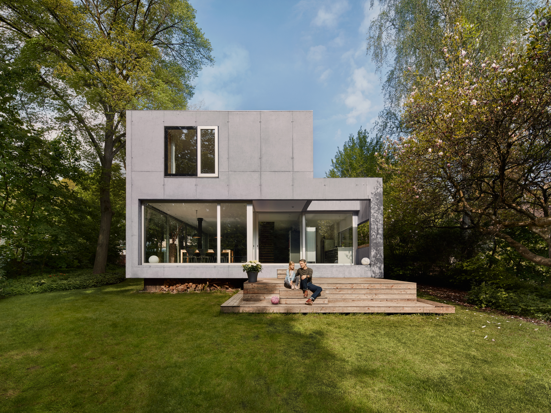 Einfamilienhaus mit Sichtbetonfassade und Glasfront, umgeben von Bäumen und gesäumt von einer Grasfläche. Auf den Stufen vor dem Haus sitzen eine erwachsene Person und ein Kind.