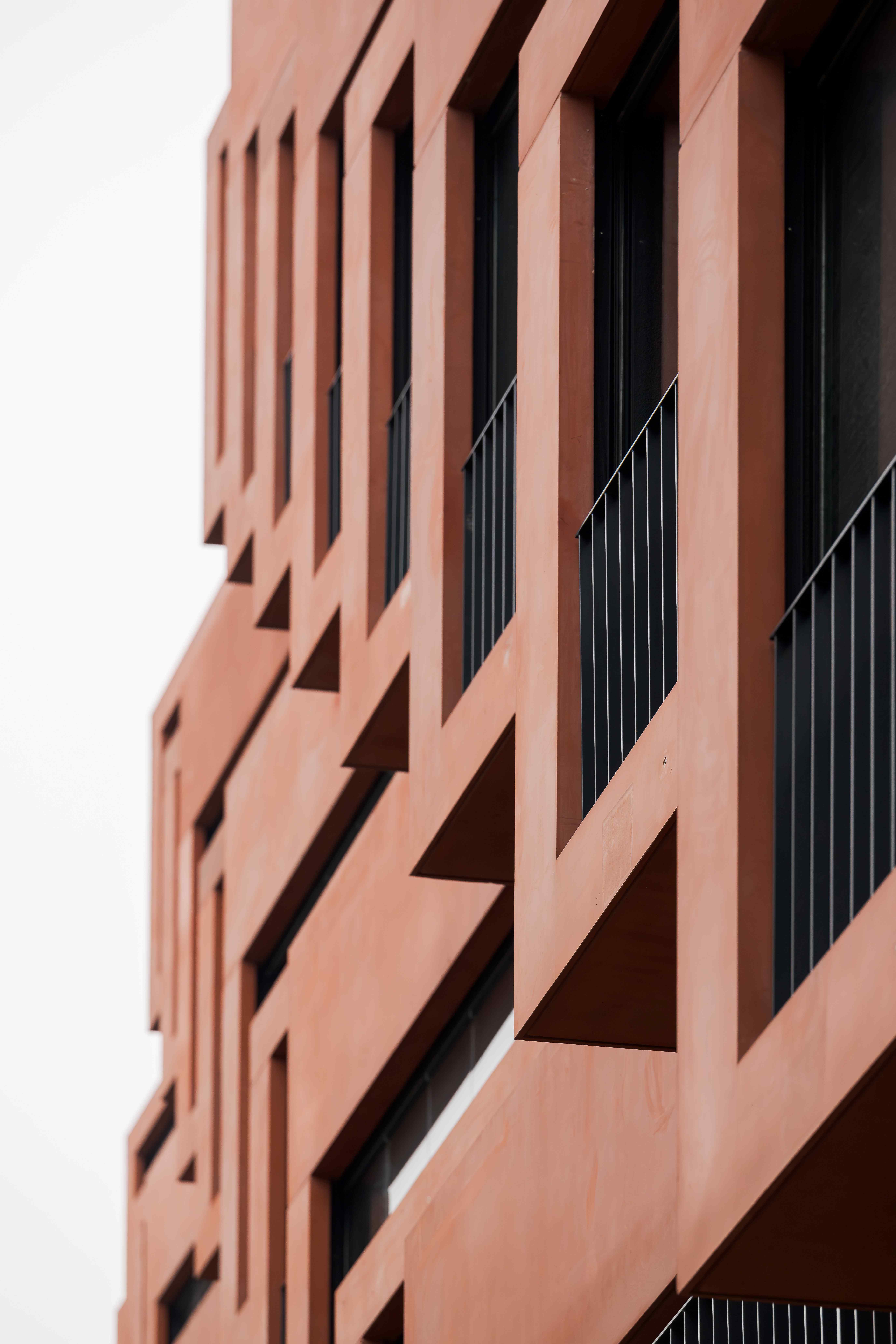 Rotes modernes Wohngebäude mit geometrischen Balkonen und schwarzen Geländern. Ansicht von unten, Fokus auf klare Linien und minimalistische Architektur.