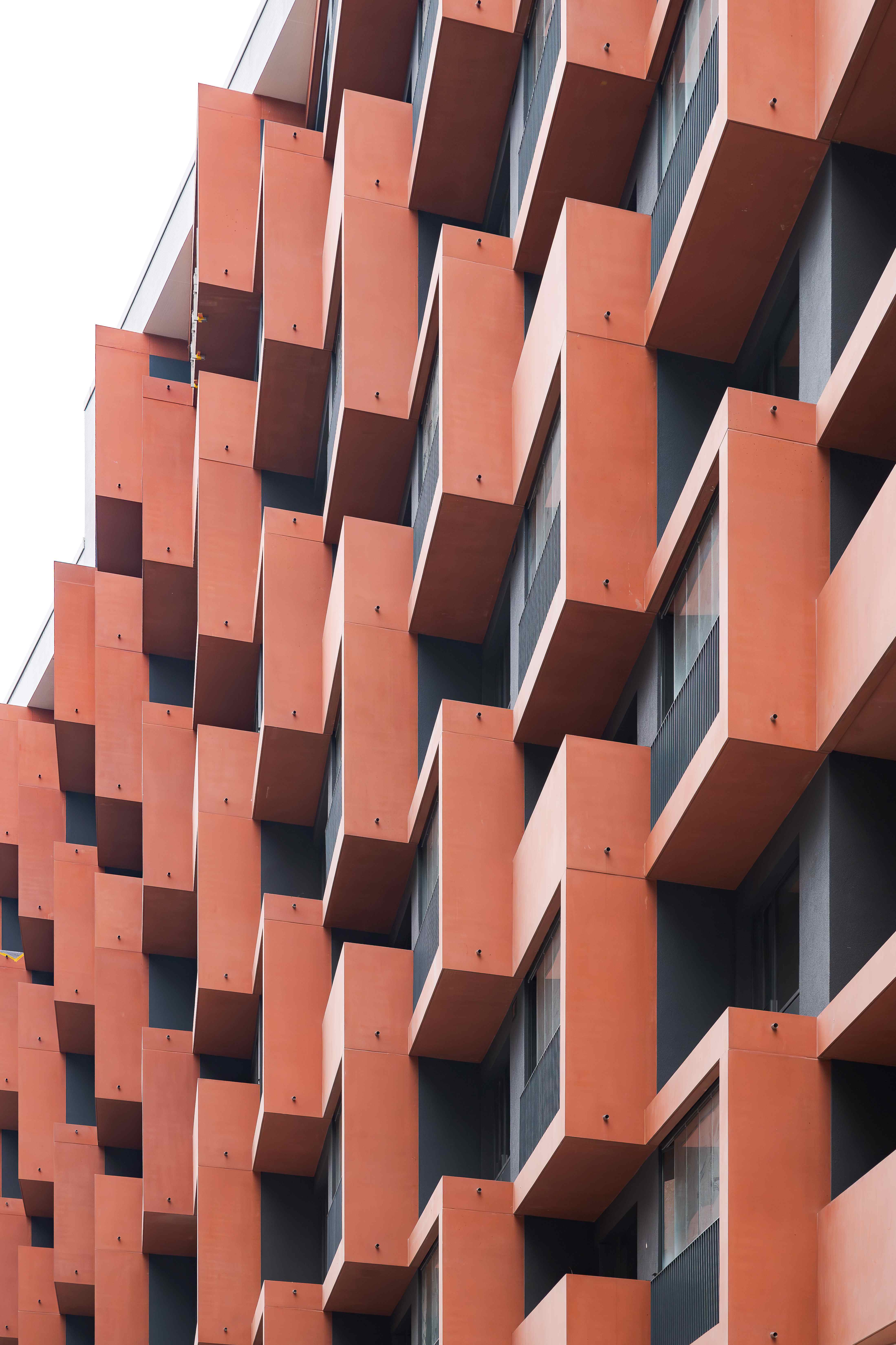 Fassade eines modernen Gebäudes mit orangefarbenen, rechteckigen Balkonelementen, die sich in einem geometrischen, versetzten Muster wiederholen; bildet ein dynamisches, architektonisches Design.