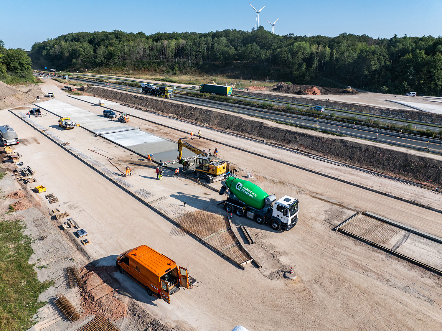 Eine im Bau befindliche Autobahn, auf der mehrere Fahrspuren betoniert werden. Ein Betonmischer von Heidelberg Materials und ein orangefarbenes Nutzfahrzeug stehen im Vordergrund, Arbeiter und Geräte sind zu sehen.