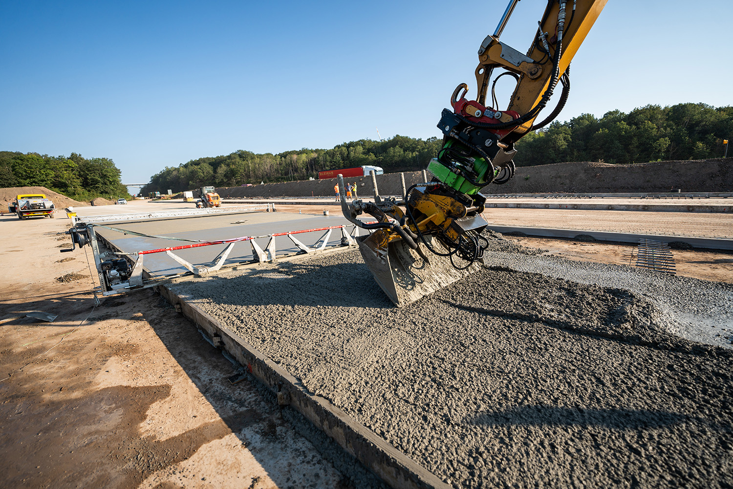 Bagger mit speziellem Anbaugerät beim Verteilen von frischem Beton auf einer Baustelle, vor dem Hintergrund unfertiger Straßenarbeiten.