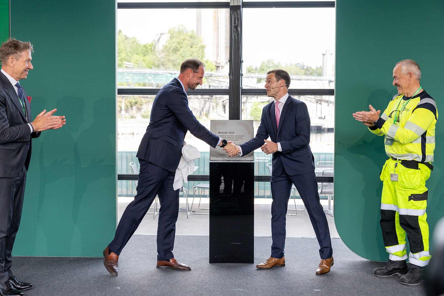 Kronprinz Haakon von Norwegen und Dr. Dominik von Achten schütteln sich in der Bildmitte die Hand vor einer schwarzen Box mit Gedenktafel aus Beton, während zwei weitere Männer – einer in Anzug (links) und einer in gelber Sicherheitskleidung (rechts) – applaudieren.