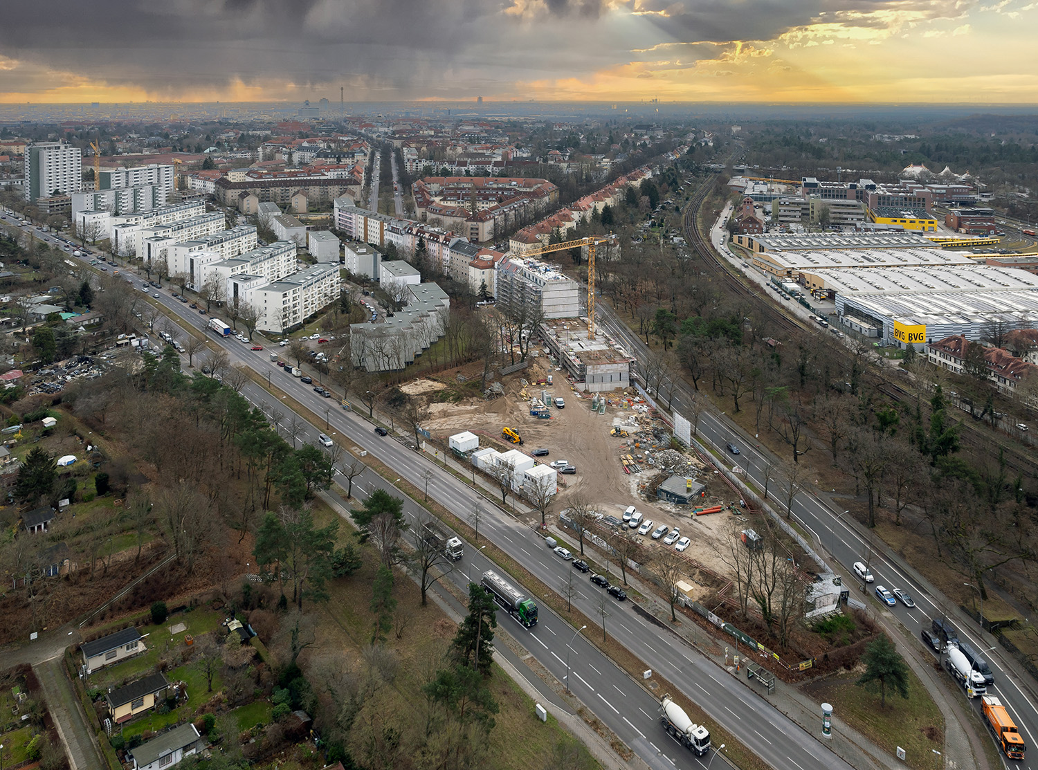 Luftaufnahme einer städtischen Baustelle in einem dicht bebauten Gebiet. Links Wohnblöcke und Grünanlagen, rechts Gewerbegebiete mit einem großen gelben Gebäudekomple. Im Zentrum die Baustelle mit Kränen und Baucontainern. Im Vordergrund verläuft eine vielbefahrene Straße, mittig durchzogen von einer Straßenbahnlinie. Wolkenbedeckter Himmel im Hintergrund.