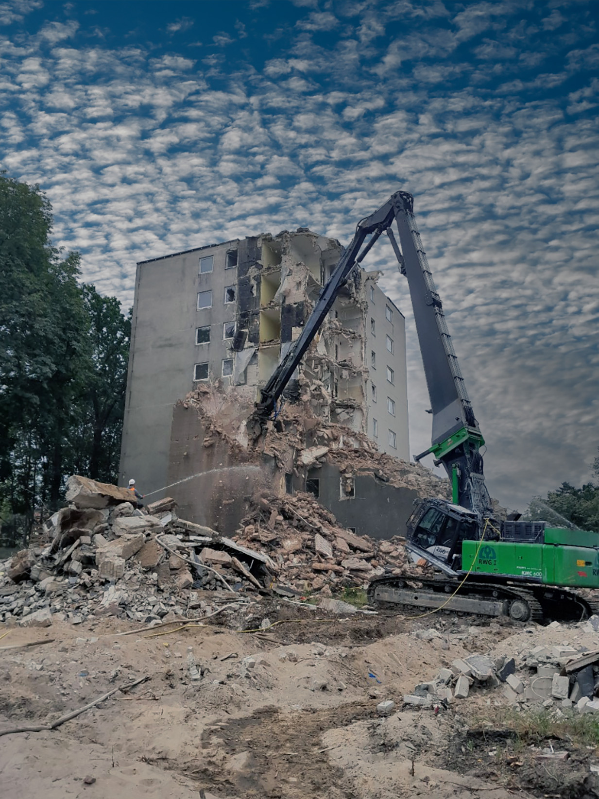 Ein Abrissbagger mit langem Arm reißt ein mehrstöckiges Wohngebäude ein. Im Hintergrund dichter Baumwuchs und ein bewölkter Himmel.