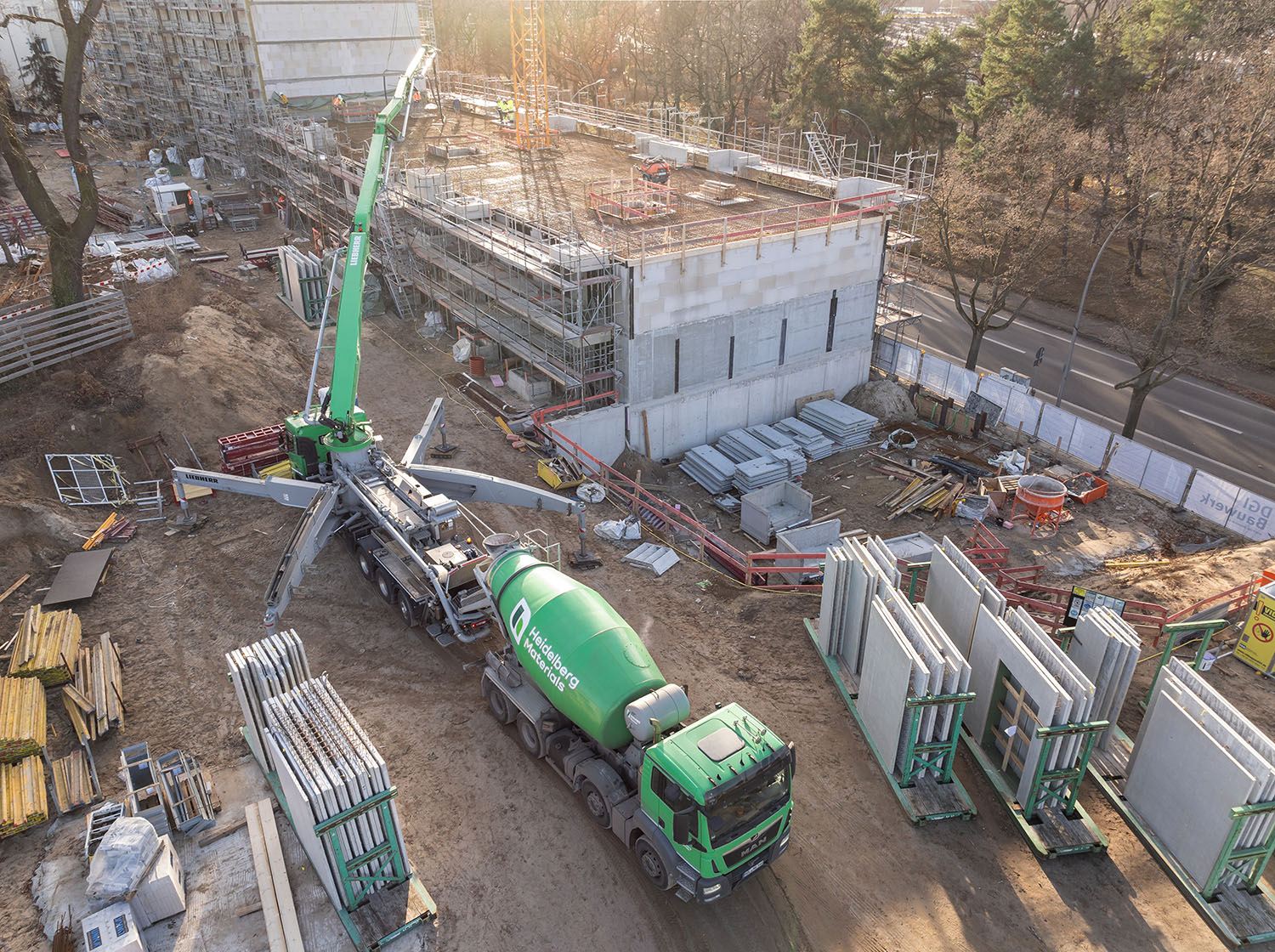 Baustelle mit halb fertiggestelltem Gebäude im Hintergrund, Betonmischer und Betonpumpe von Heidelberg Materials im Vordergrund, sowie gestapelte Wandmodule rechts. Baumaterialien und Baugeräte links und unten verteilt. Baustelle von oben bei Tageslicht mit umliegenden Bäumen.