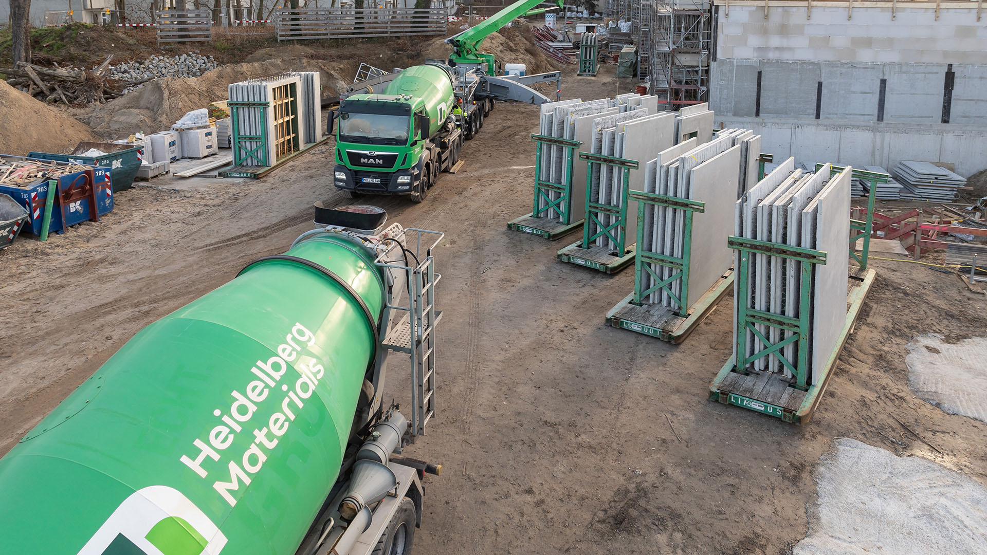 Grüne Betonmischer der Marke Heidelberg Materials auf einer Baustelle, umgeben von Betonfertigteilen auf Paletten; im Hintergrund sind Baugeräte, ein Kran und ein teilweise errichtetes Gebäude zu sehen.
