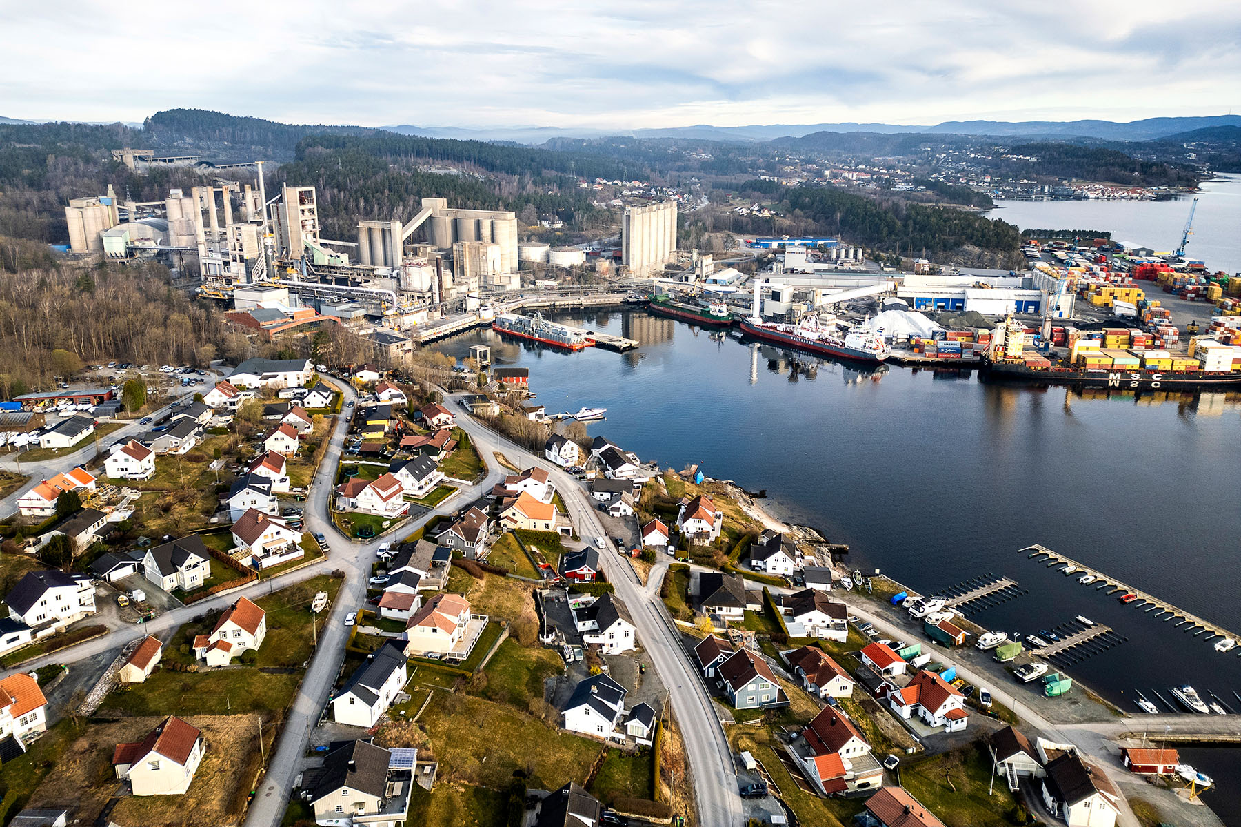 Luftaufnahme von Brevik in Norwegen mit dicht bebauten Wohnhäusern im Vordergrund, großem Zementwerk auf der linken Seite sowie einem Containerhafen rechts am Wasser. Im Hintergrund bewaldete Hügel und ein See.