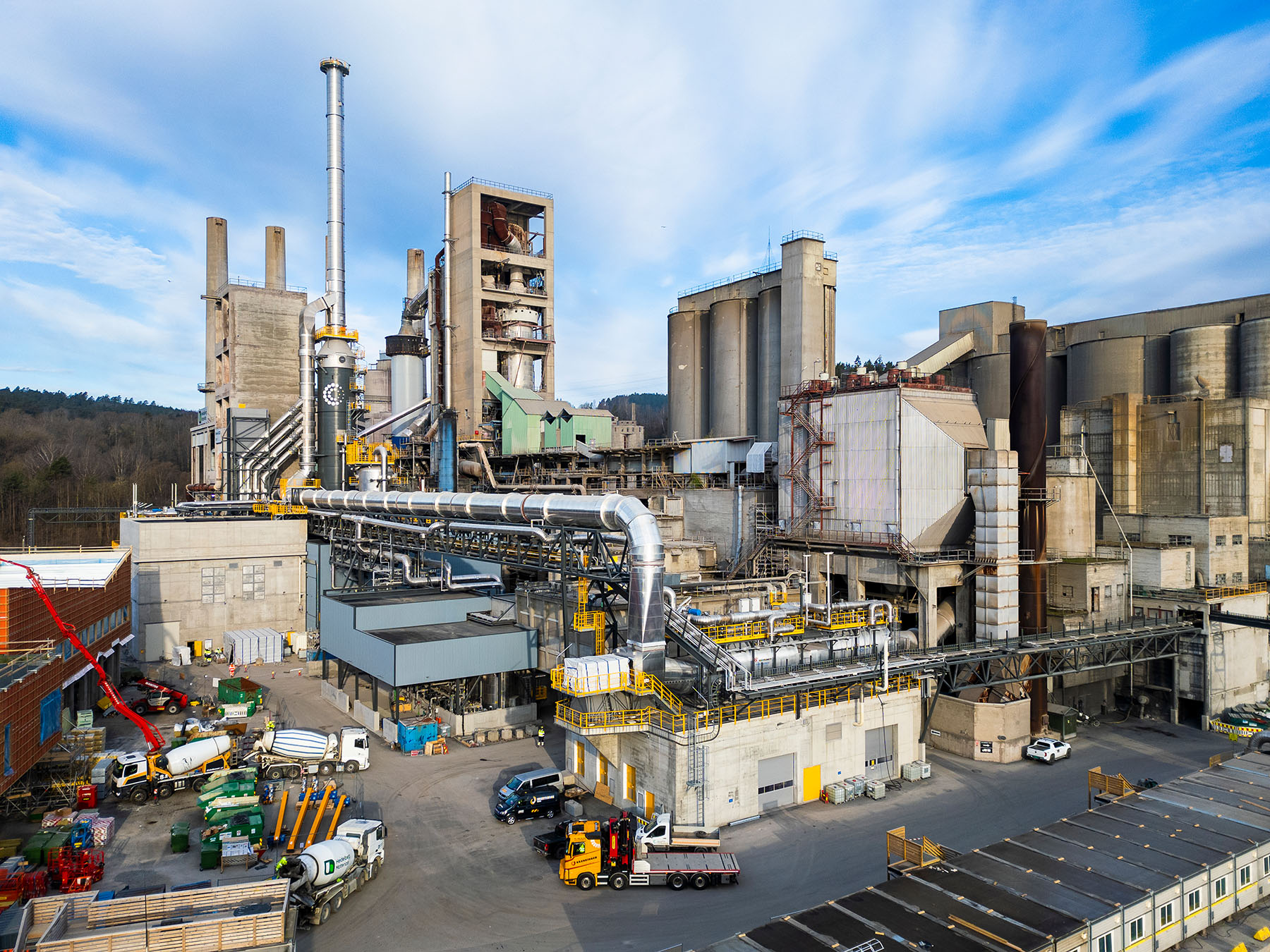 Zementwerk mit mehreren großen Silos, Rohrleitungen und CO2-Abscheideanlage in zentraler Position; im Vordergrund LKWs, Container und Baumaschinen; umliegende grüne Landschaft im Hintergrund unter bewölktem Himmel.