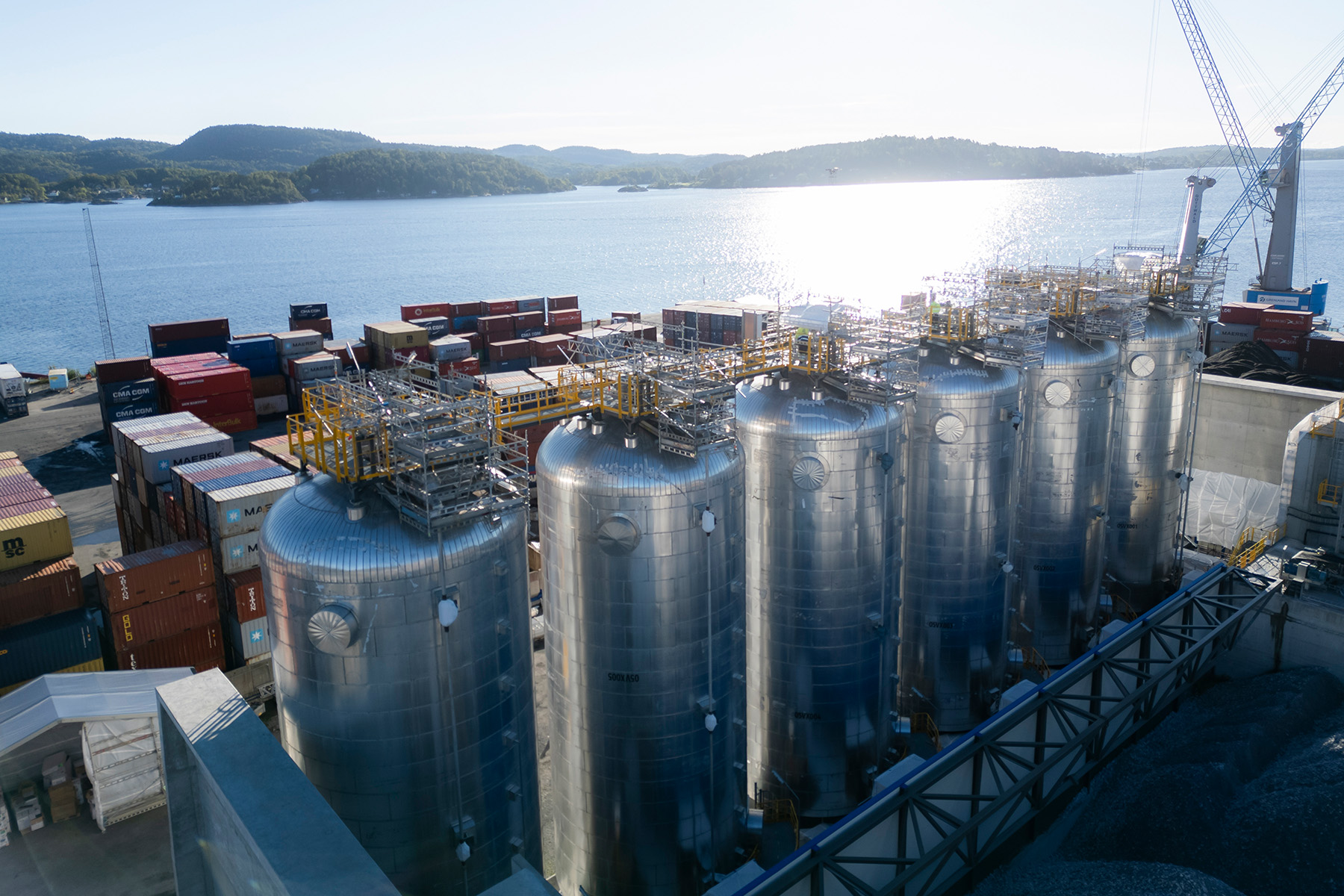 Reihe großer, silberner CO2-Silos in einer Hafenanlage, umgeben von Containern und Kränen; im Hintergrund Wasser und bewaldete Hügel bei hellem Sonnenlicht.