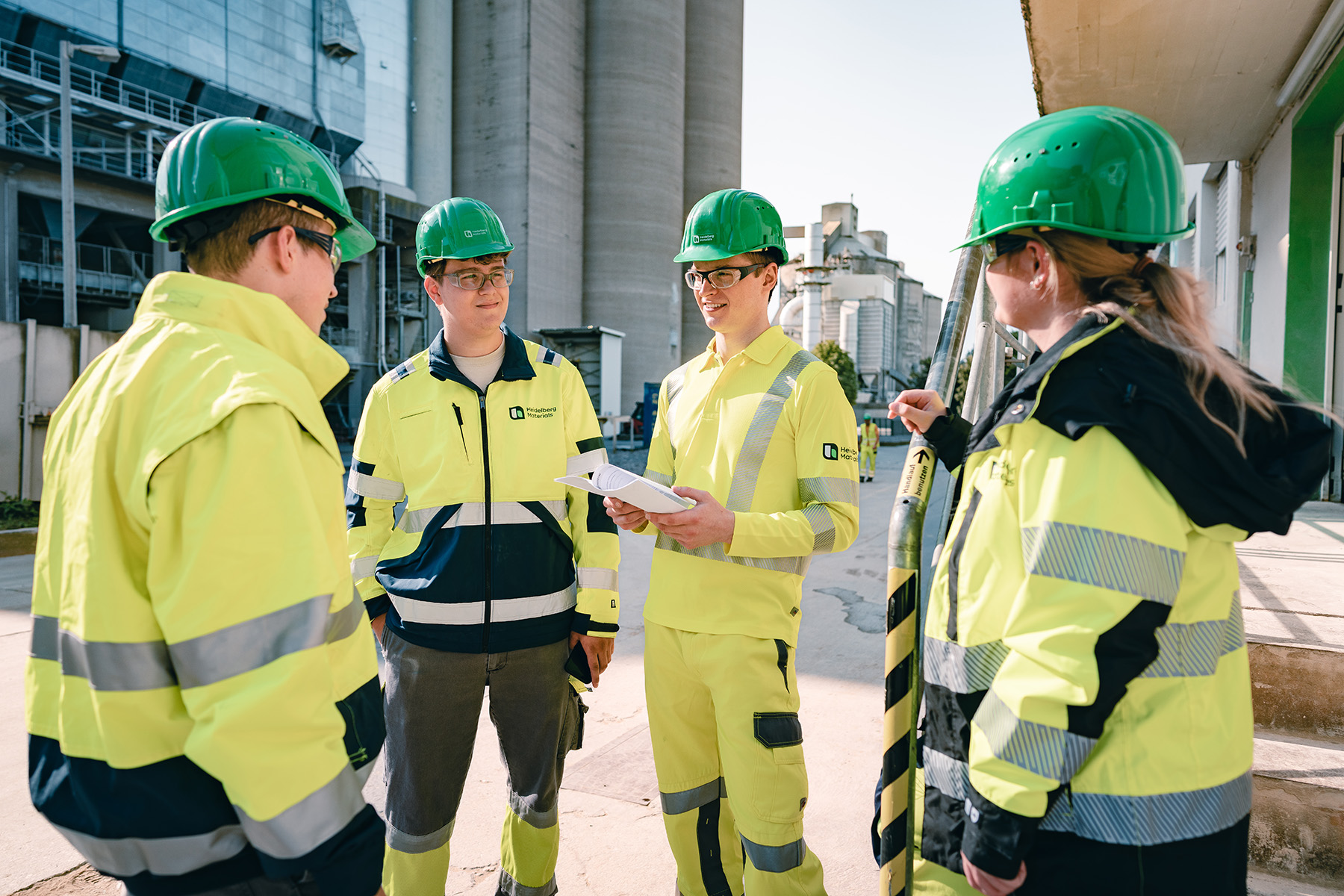 Vier Personen von Heidelberg Materials in gelber PSA und grünen Helmen stehen in einem Zementwerk.