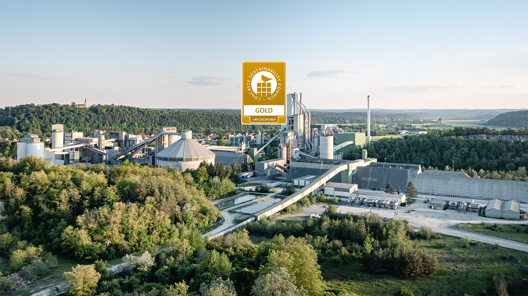Luftaufnahme eines des Zementwerks Burglengenfeld inmitten einer grünen Landschaft mit Wald und Wiesen. Zentral über dem Werk wird das CSC Zertifikat gezeigt.