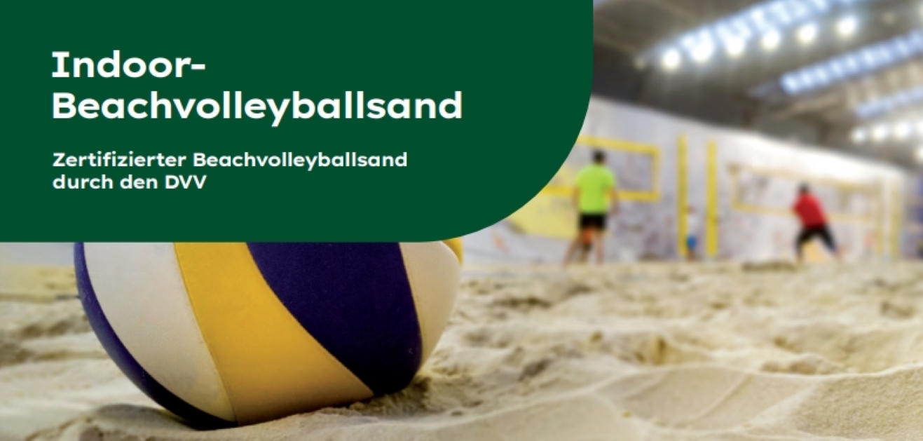 Nahaufnahme eines Volleyballs im Sand in einer Indoor-Beachvolleyballhalle. Im Hintergrund sind unscharf zwei Personen beim Spielen vor einem gelben Netz zu sehen. Links oben befindet sich ein dunkelgrünes Banner mit der Aufschrift „Indoor-Beachvolleyballsand – Zertifizierter Beachvolleyballsand durch den DVV“.