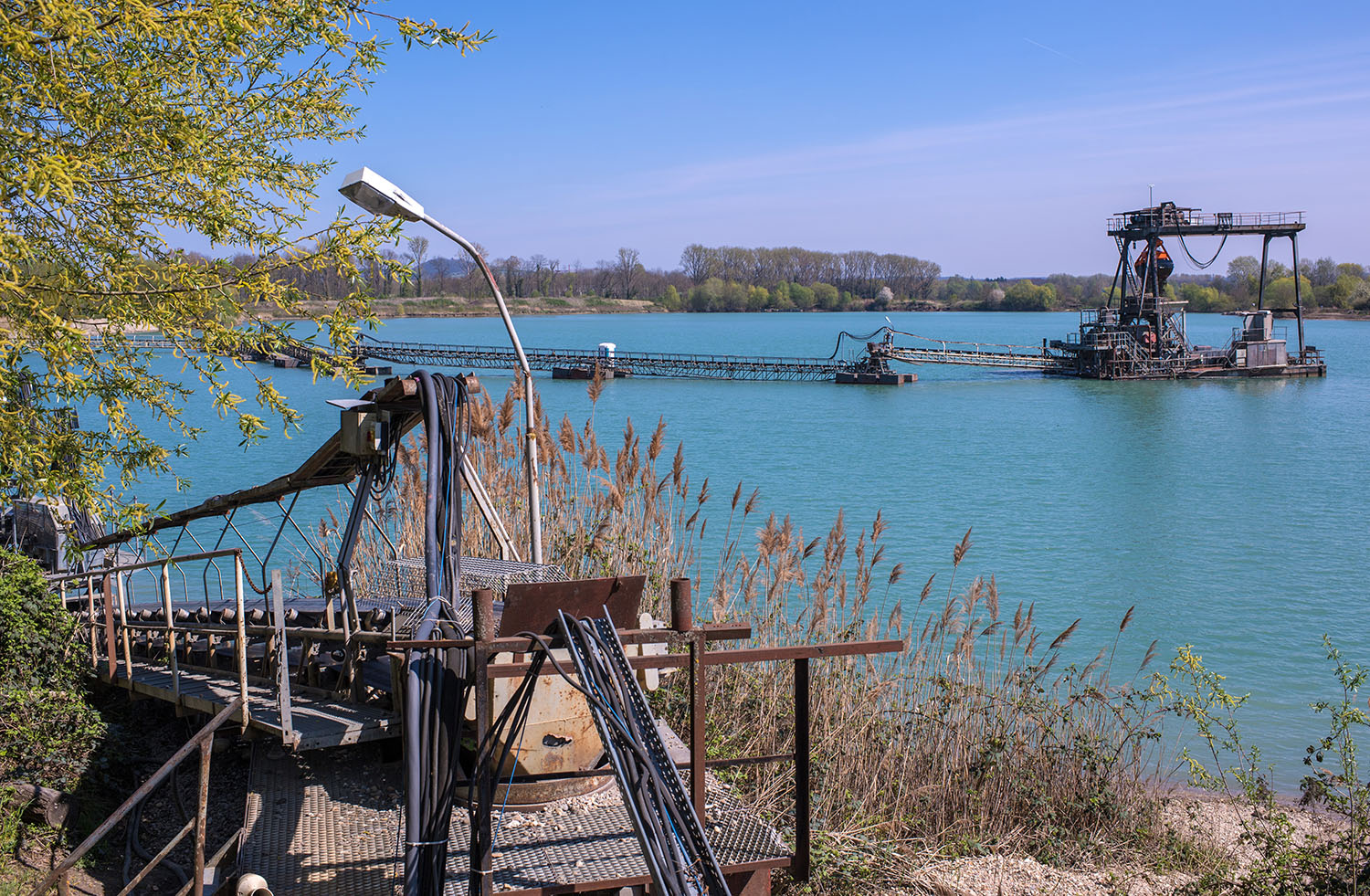 Sand- und Kiesabbau an einem See: Im Vordergrund ein Metallsteg mit Rohren und Geländer, umgeben von Schilf. Im Hintergrund eine Pontonplattform mit Förderband und Konstruktion für Kies- oder Sandabbau auf dem Wasser. Klarer Himmel und Bäume entlang des Seeufers.