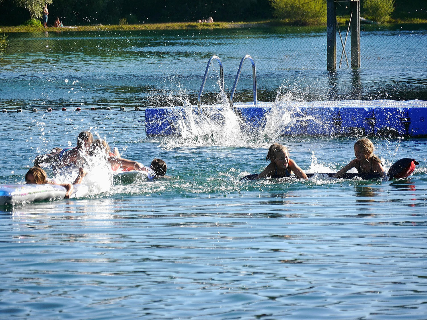 Kinder schwimmen in einem See. Im Hintergrund befinden sich ein Steg, eine Leiter und Bojen, die den Schwimmbereich abgrenzen. Spritzendes Wasser zeigt aktive Bewegung.