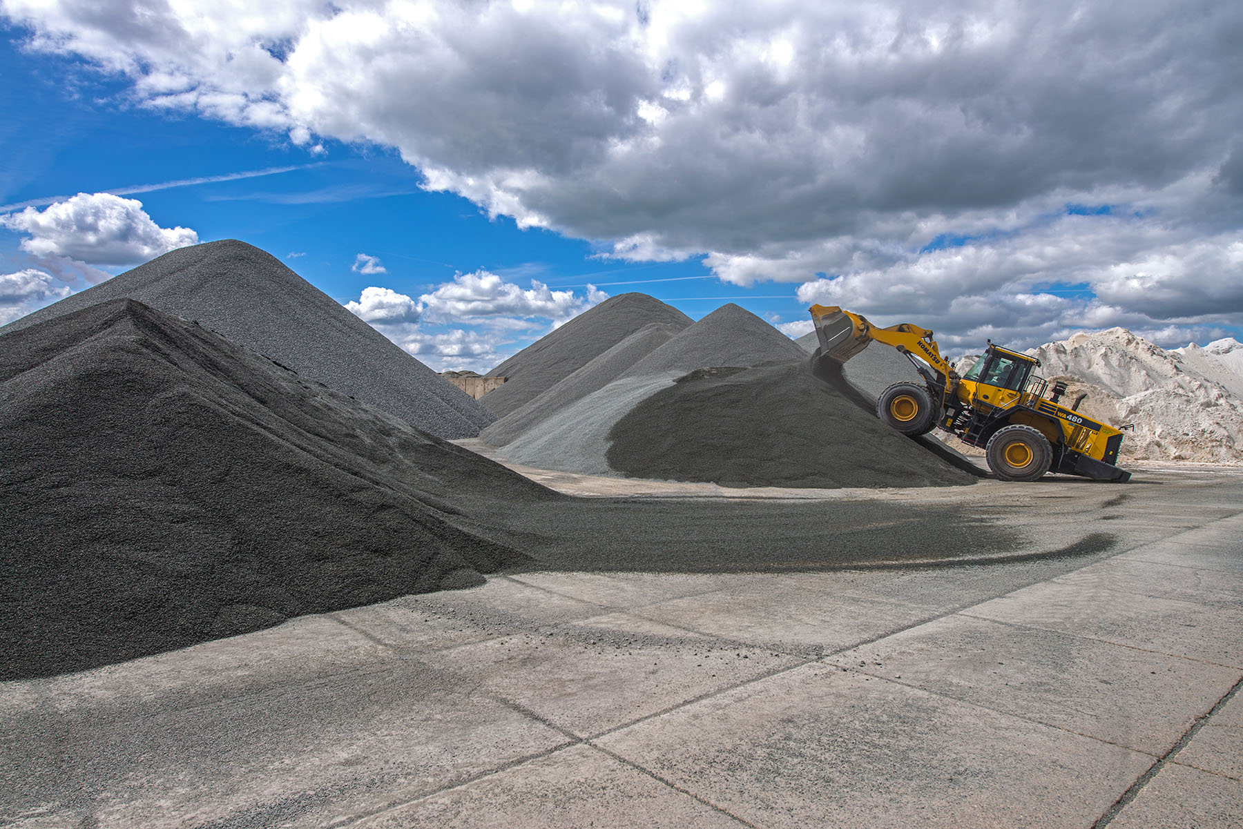 Gelber Radlader an einer grauen Kieshalde im Kieswerk, umgeben von mehreren Kieshaufen; Himmel bedeckt mit Wolken.