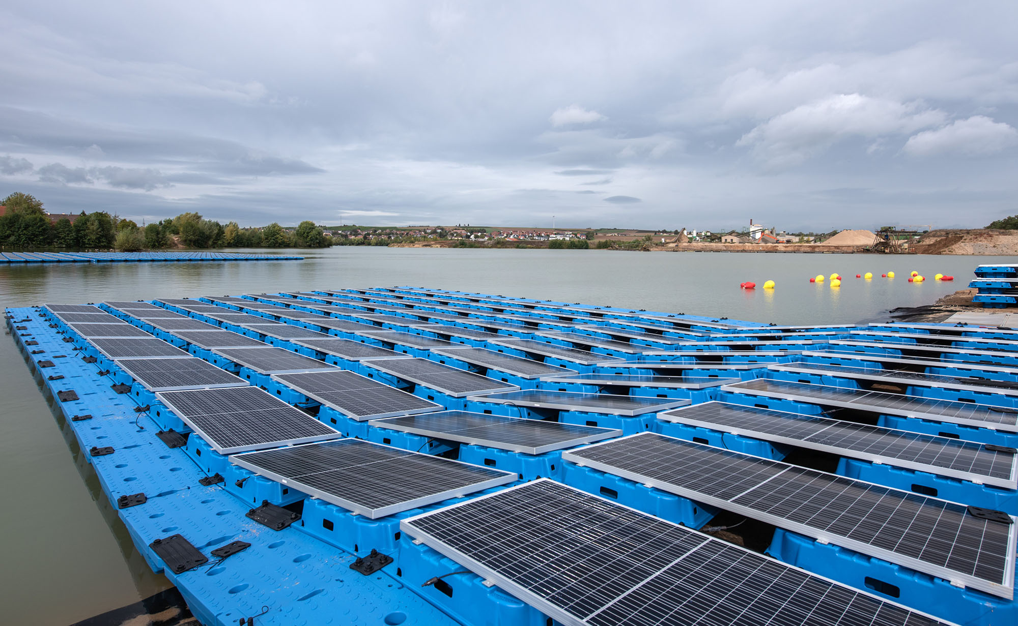 Schwimmende Solaranlage auf einem Baggersee, bestehend aus blauen Plattformen mit montierten Solarmodulen. Im Hintergrund sind Bäume, Industriebauten und gelbe Bojen sichtbar. Der Himmel ist bewölkt.