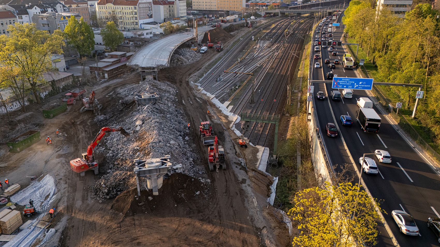 Luftaufnahme einer Baustelle neben einer Autobahn über Eisenbahnschienen in einer Stadt. Im linken Bereich ist ein großer Erdhügel mit Baggern und Bauarbeitern zu sehen. Rechts verlaufen dicht befahrene Fahrspuren der Autobahn parallel zu Gleisen. Im Hintergrund sind Stadtgebäude und Bäume im Abendlicht erkennbar.