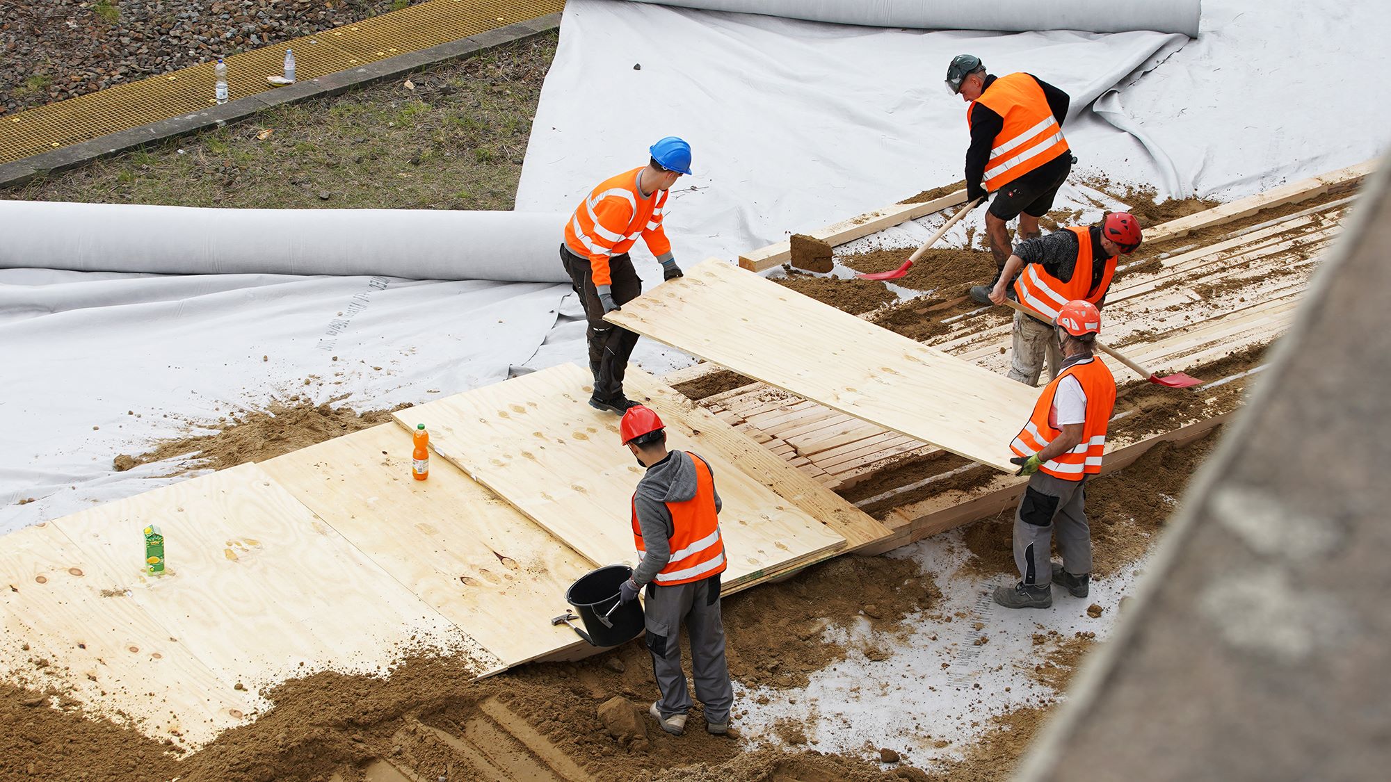 Fünf Bauarbeiter in orangefarbenen Sicherheitswesten verlegen Holzplatten über die mit weißer Plane abgedeckten Gleise; zwei Arbeiter halten eine Platte, die sie positionieren.