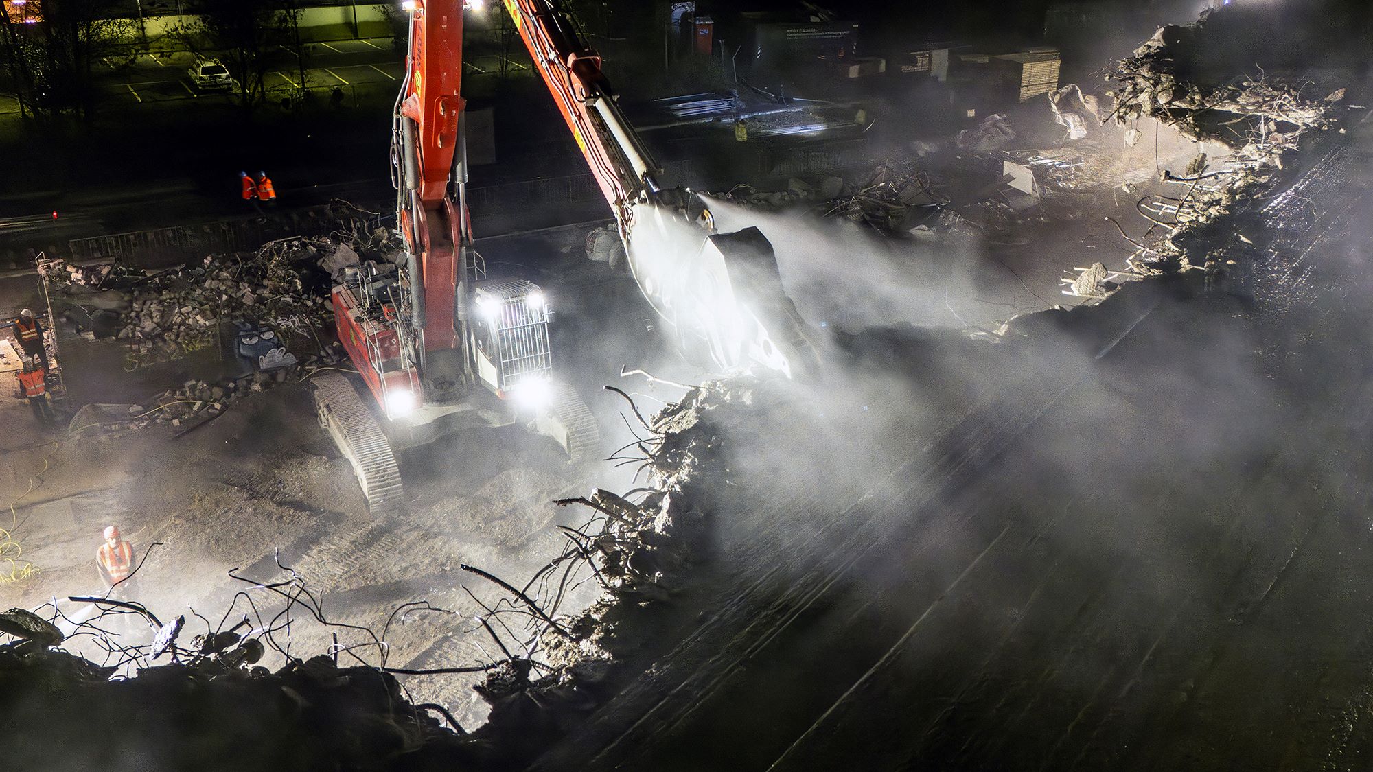 Nächtliche Abrissarbeiten an einer Brücke: Ein großer Bagger reißt Betonstrukturen ein, während Staub und Schutt in die Luft wirbeln. Arbeiter in Schutzkleidung und Warnwesten stehen im Hintergrund. Baustellenbeleuchtung erhellt die Szene.