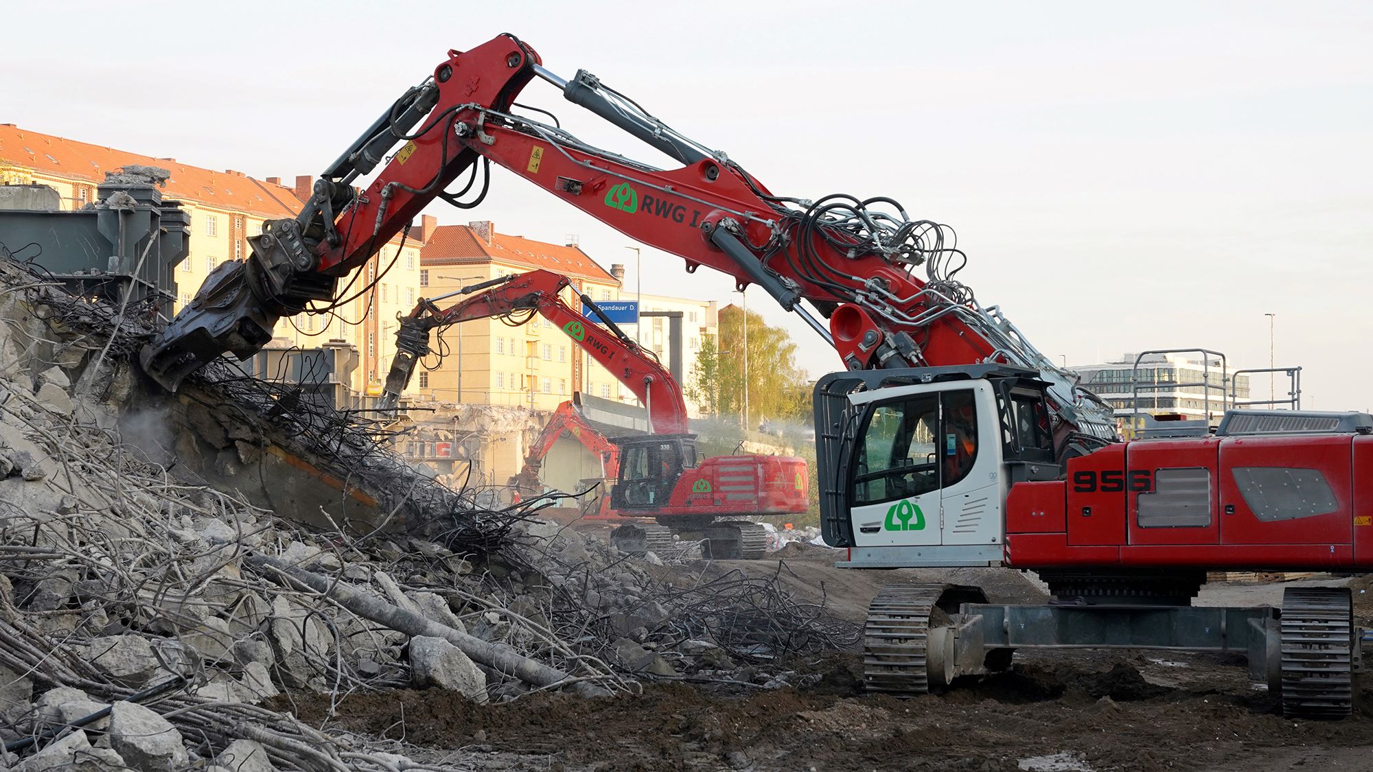 Zwei rot-weiße Abrissbagger entfernen Trümmer einer abgerissenen Brücke auf einer Baustelle. Im Hintergrund sind Gebäude sichtbar.