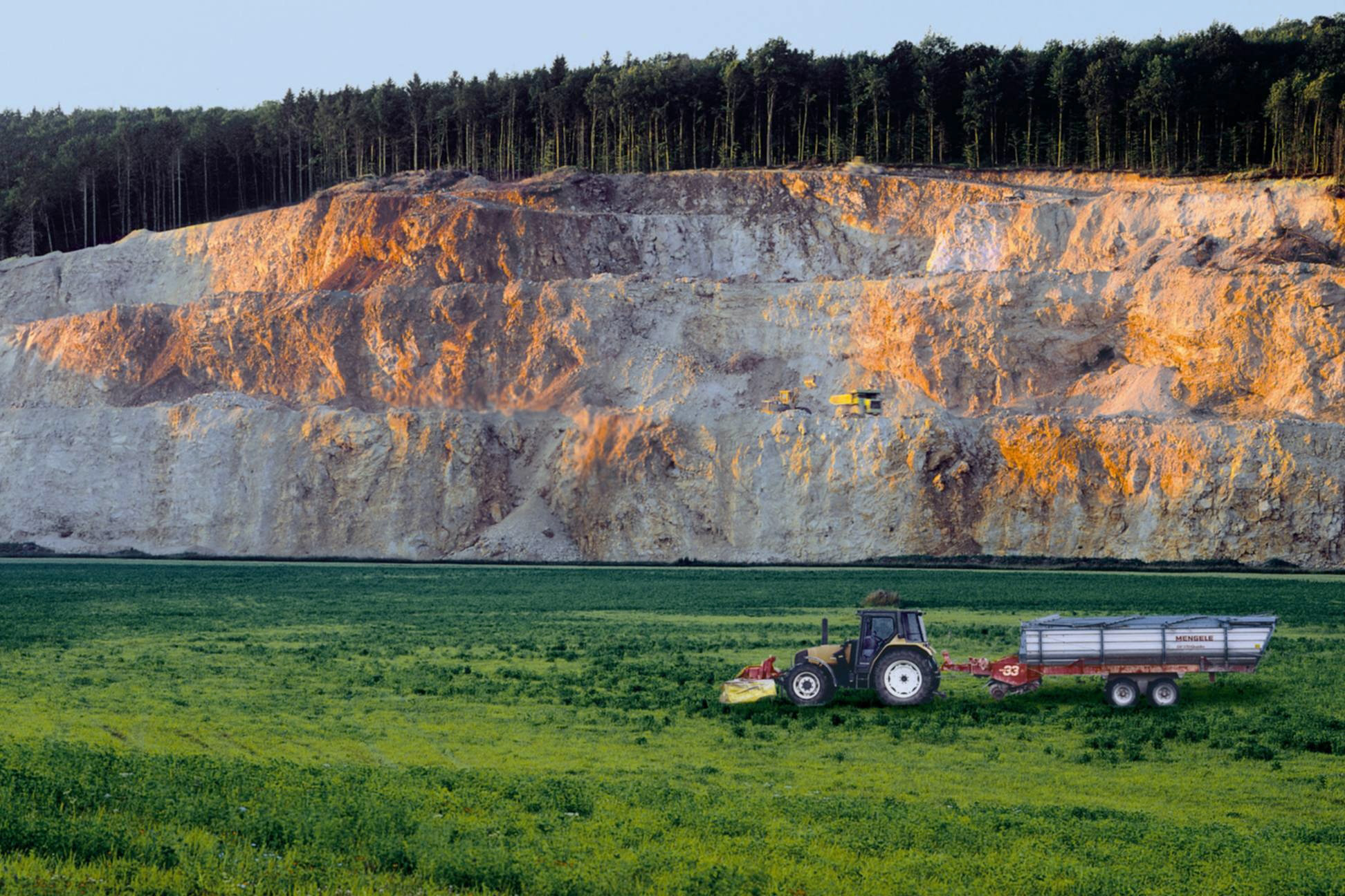 Ein Traktor mit einem Anhänger steht auf einer grünen Wiese im Vordergrund, während im Hintergrund eine große, steil abfallende Kalksteinformation zu sehen ist. Oben auf der Formation befindet sich ein dichter Wald. Die Abendsonne beleuchtet die Felswand teils orange-golden.