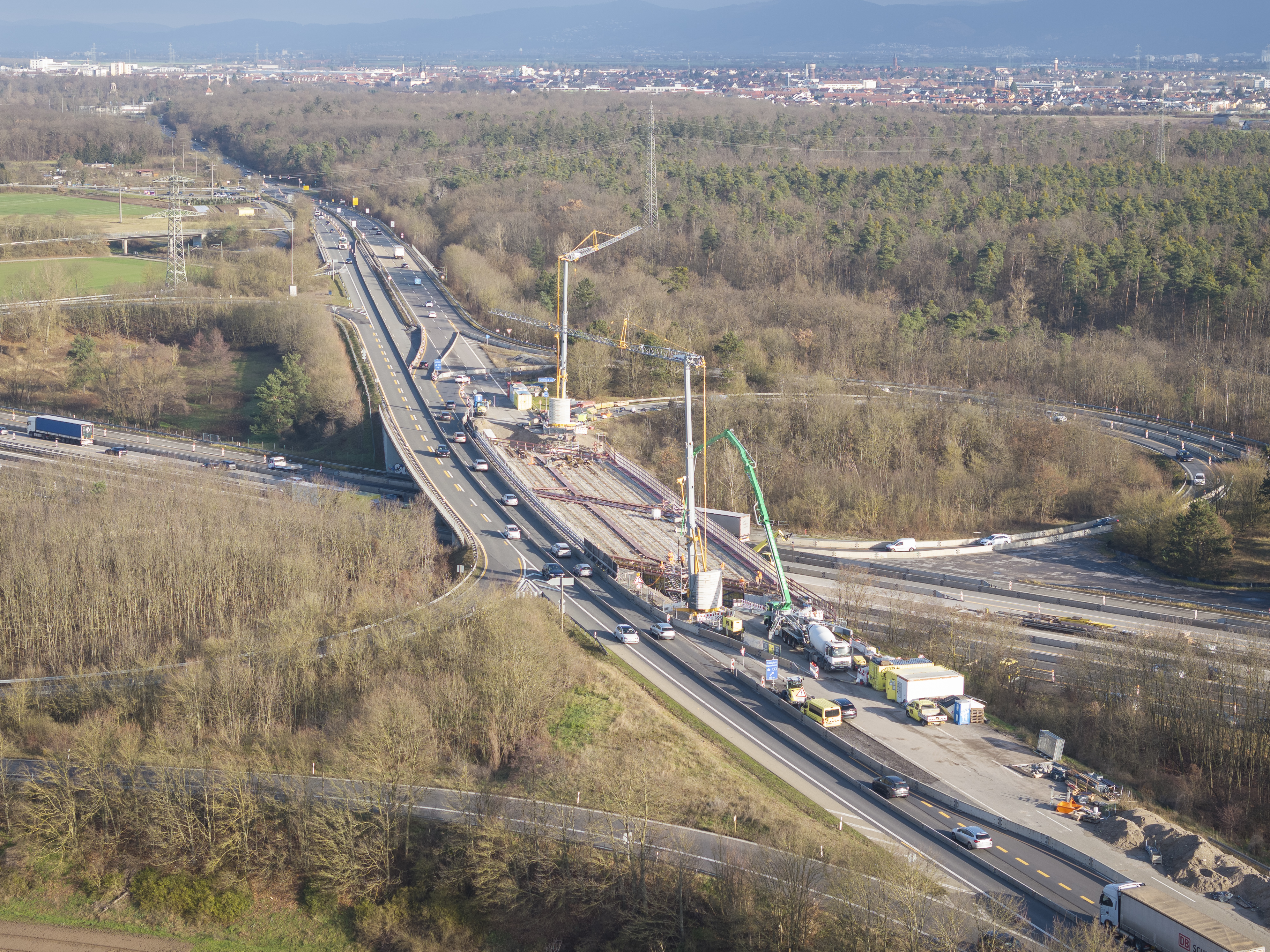 Luftaufnahme einer Autobahnbaustelle: Ein mehrspuriger Autobahnabschnitt mit laufenden Brückenarbeiten in der Bildmitte. Zwei große Kräne stehen auf der Baustelle, umgeben von Fahrzeugen und Baumaschinen. Verkehr fließt rechts und links um die Baustelle herum. Im Hintergrund sind Wälder, landwirtschaftliche Flächen und eine Stadt zu sehen.
