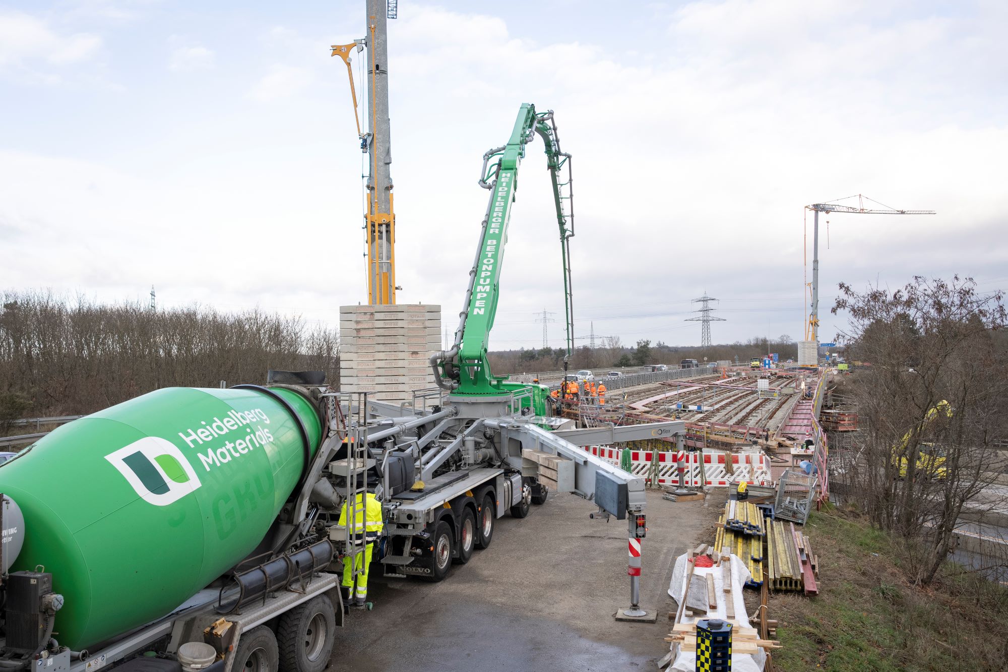 Betonmischer und Betonpumpe vorne links, Arbeiter mit Warnweste in der Mitte, Baustelle für eine Brücke im Hintergrund mit Kränen und Bahngleisen rechts.