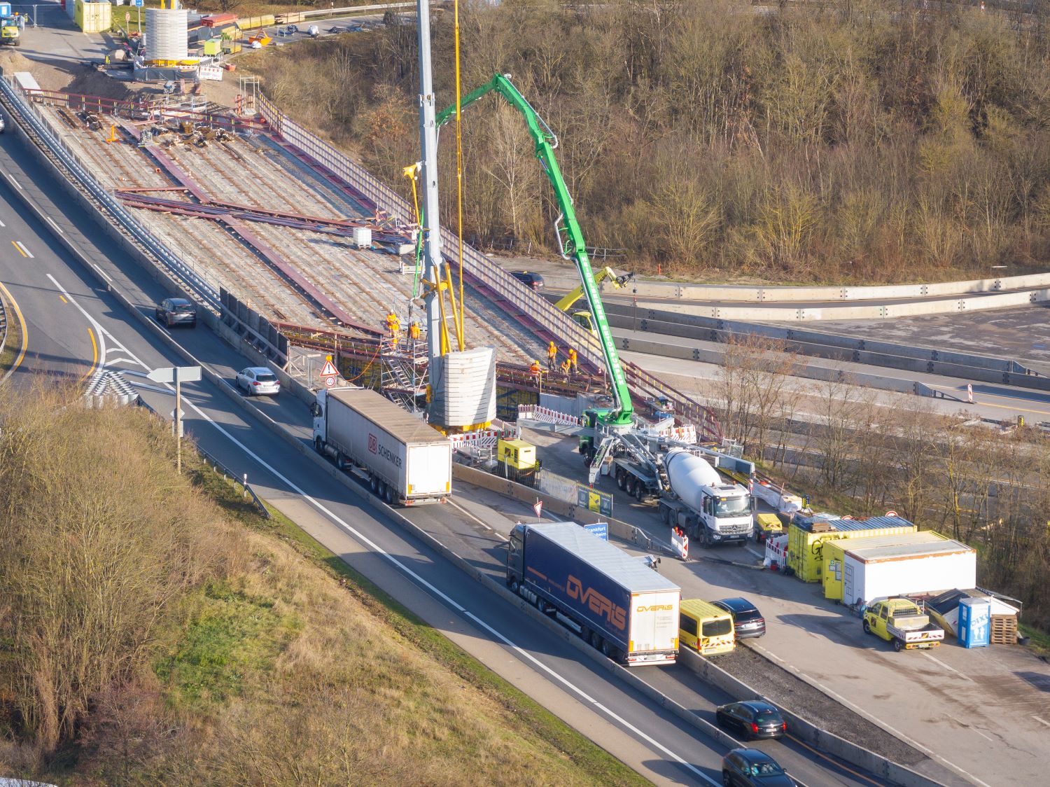 Luftaufnahme einer Baustelle auf einer Autobahnbrücke; mehrere Baustellenfahrzeuge, Betonpumpen und Arbeiter im mittleren Bereich; zwei Fahrspuren links und rechts, teilweise durch Lkws und Verkehr blockiert; umliegende Vegetation und Straßenknoten erkennbar.