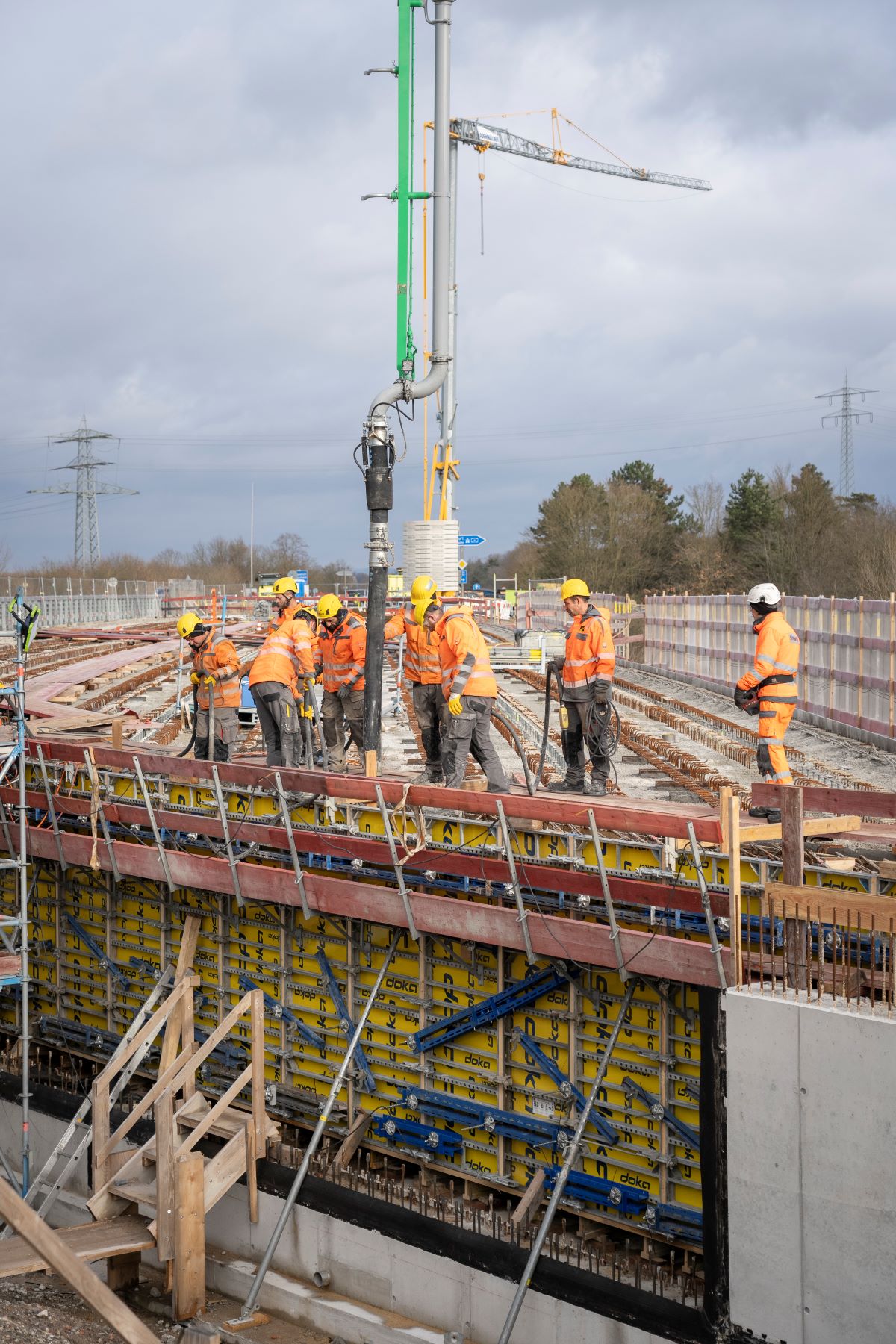 Bauarbeiter in orangefarbener Sicherheitskleidung arbeiten auf einer Brückenbaustelle. Sichtbar sind eine Betonpumpe, die Beton auf die Brücke gießt, und ein Kran im Hintergrund. Umgeben von freiem Himmel und Strommasten in der Ferne.
