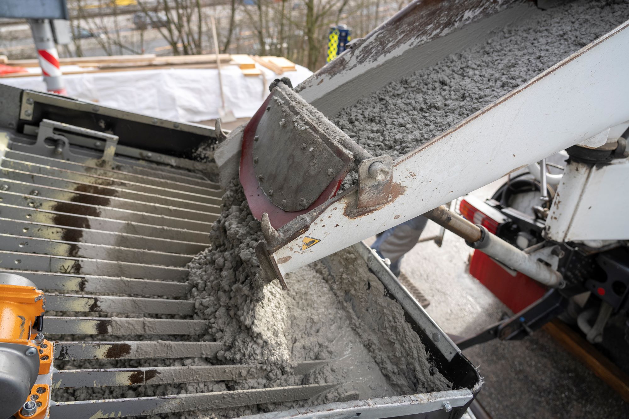 Beton fließt aus einer Betonpumpe auf eine geriffelte Stahlplattform. Im Hintergrund sind geländerartige Strukturen sichtbar.