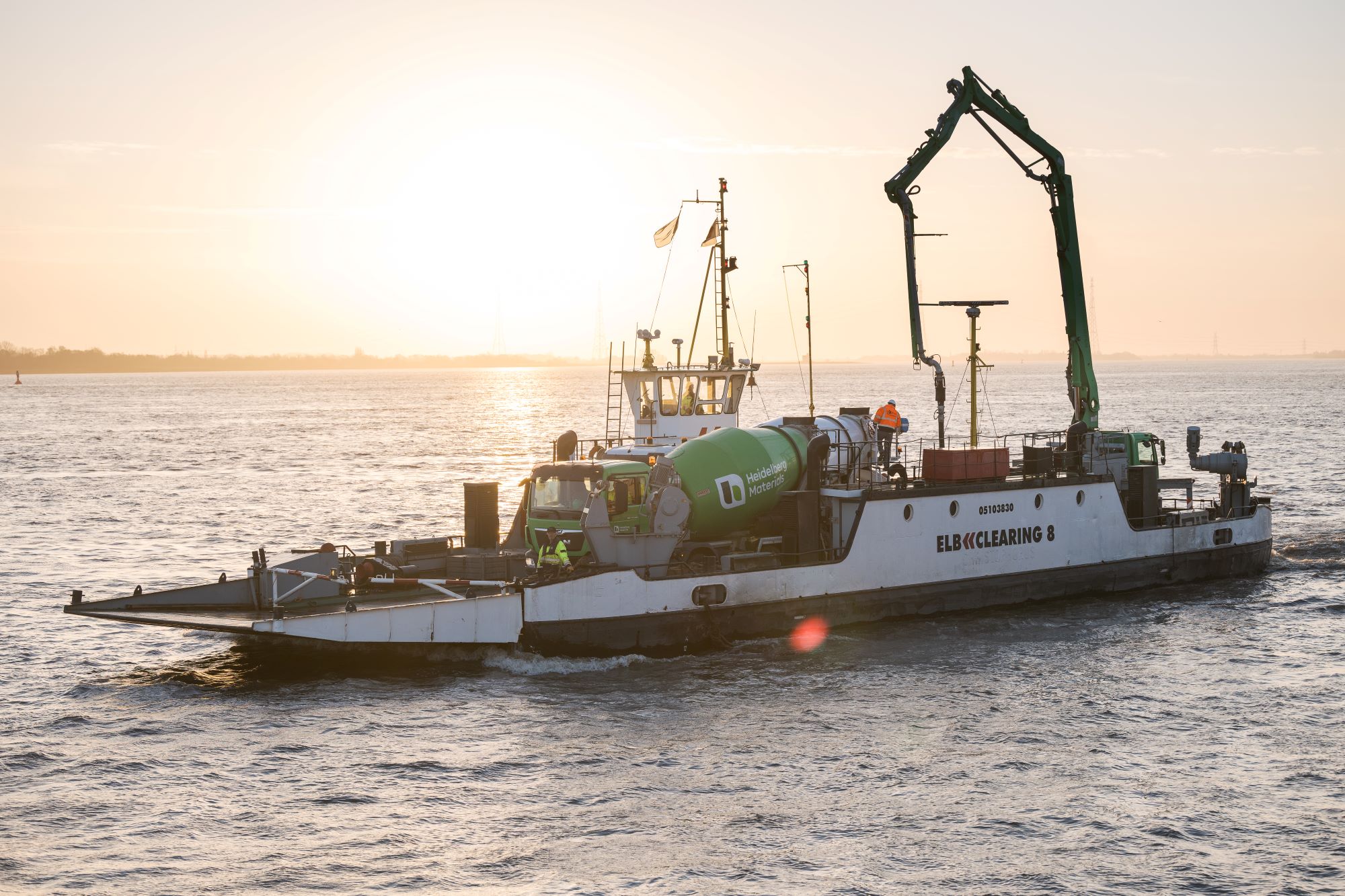 Fähre mit Betonmischer auf Deck fährt über das Wasser bei Sonnenuntergang, das Wasser reflektiert das Licht; im Hintergrund schwache Konturen einer Uferlinie sichtbar.
