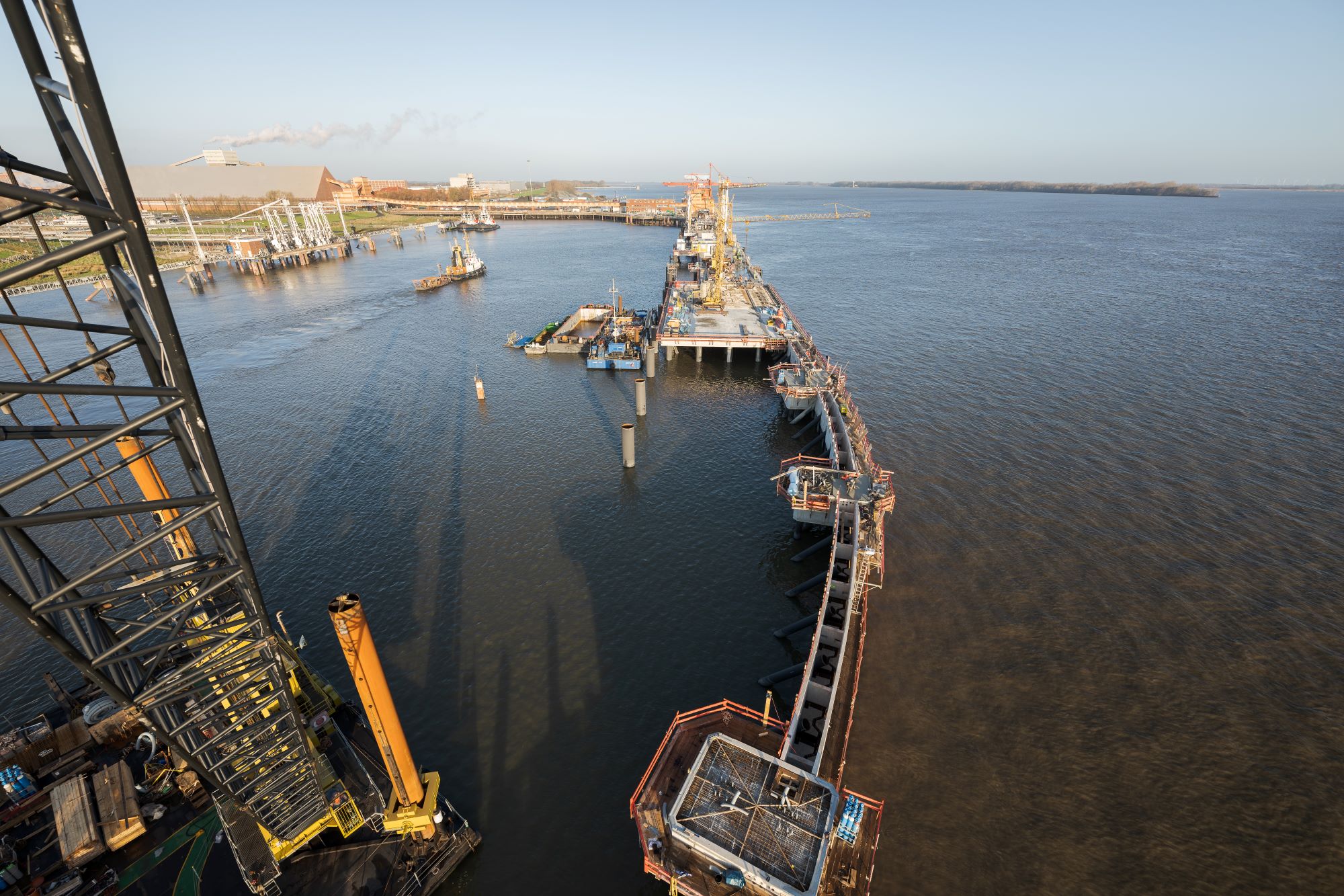 Luftaufnahme eines Hafenbauprojekts: Ein langer Pier ragt diagonal von unten rechts nach oben mitte ins Wasser. Baustellenmaterial und Kräne sind entlang des Piers verteilt. Ein Schlepper befindet sich links neben dem Pier. Im Hintergrund sind ein weiteres Ufer und eine industrielle Landschaft sichtbar.