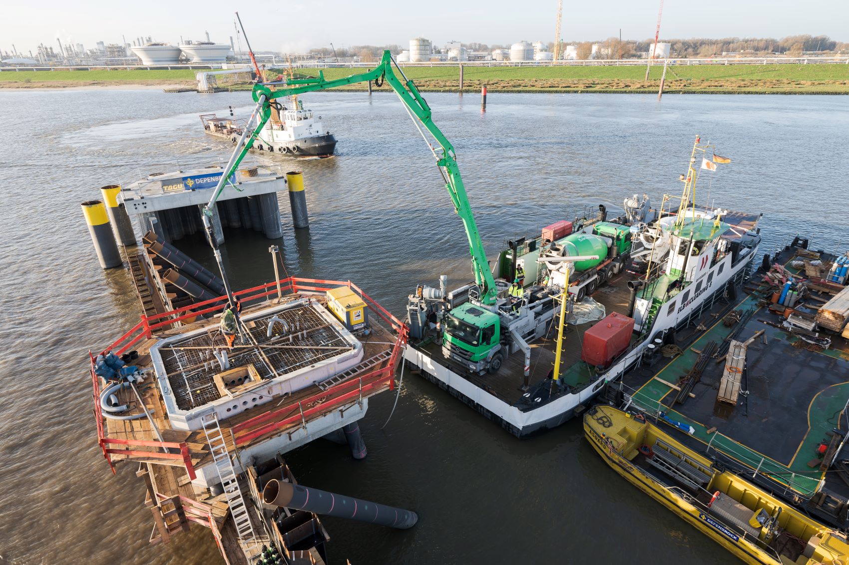 Eine Baustelle auf dem Wasser, bei der eine grüne Betonpumpe von einem Schiff aus Beton auf eine kreisförmige Plattform links pumpt. Im Hintergrund weitere Schiffe und ein Ufer mit Industriegebäuden.
