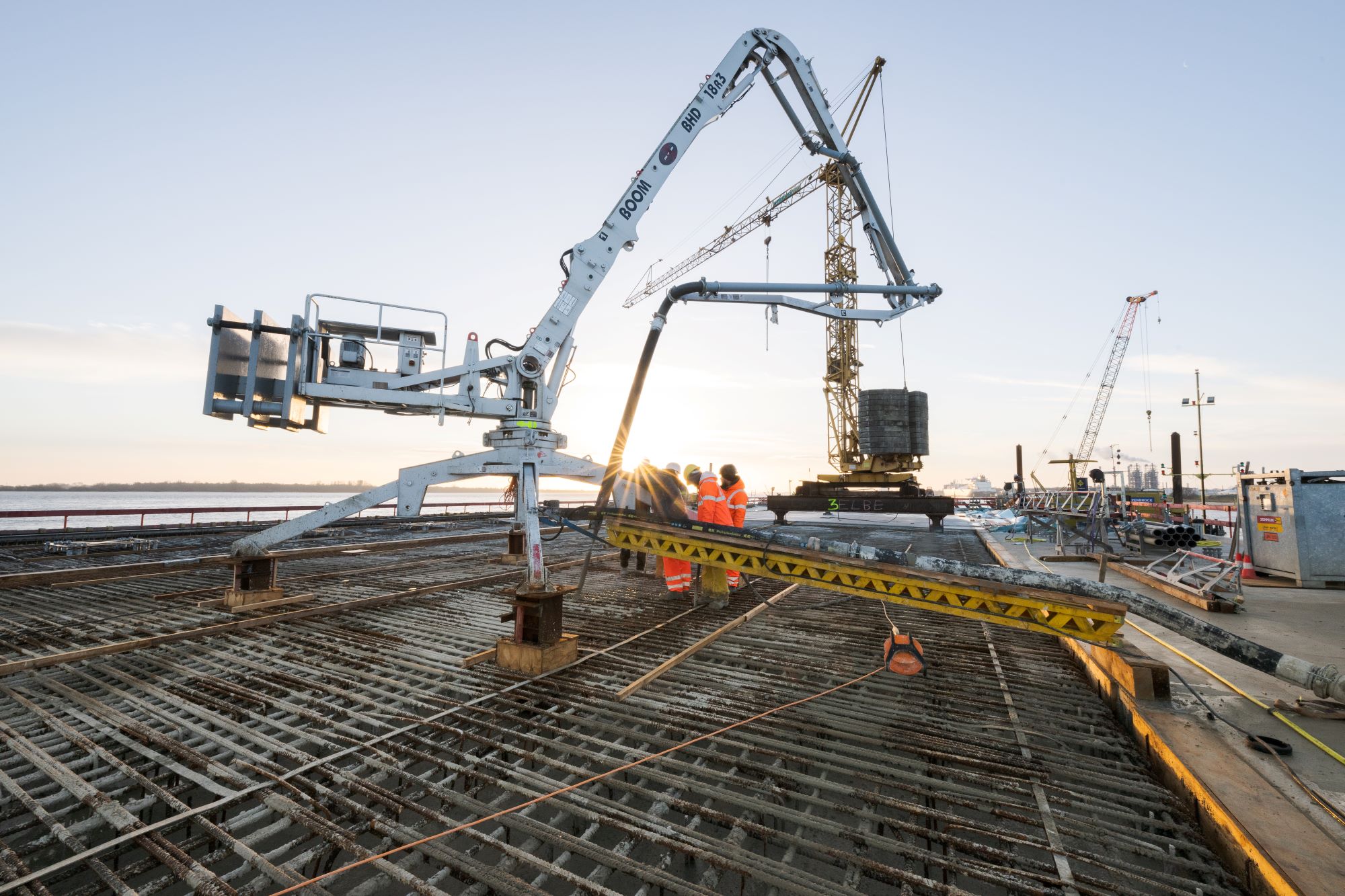 Baustelle mit Betonpumpe und Bauarbeitern, die Beton auf eine Stahlbewehrung gießen; im Hintergrund Kräne und Sonnenaufgang, zentrale Betonierarbeiten auf Brückenfundament.