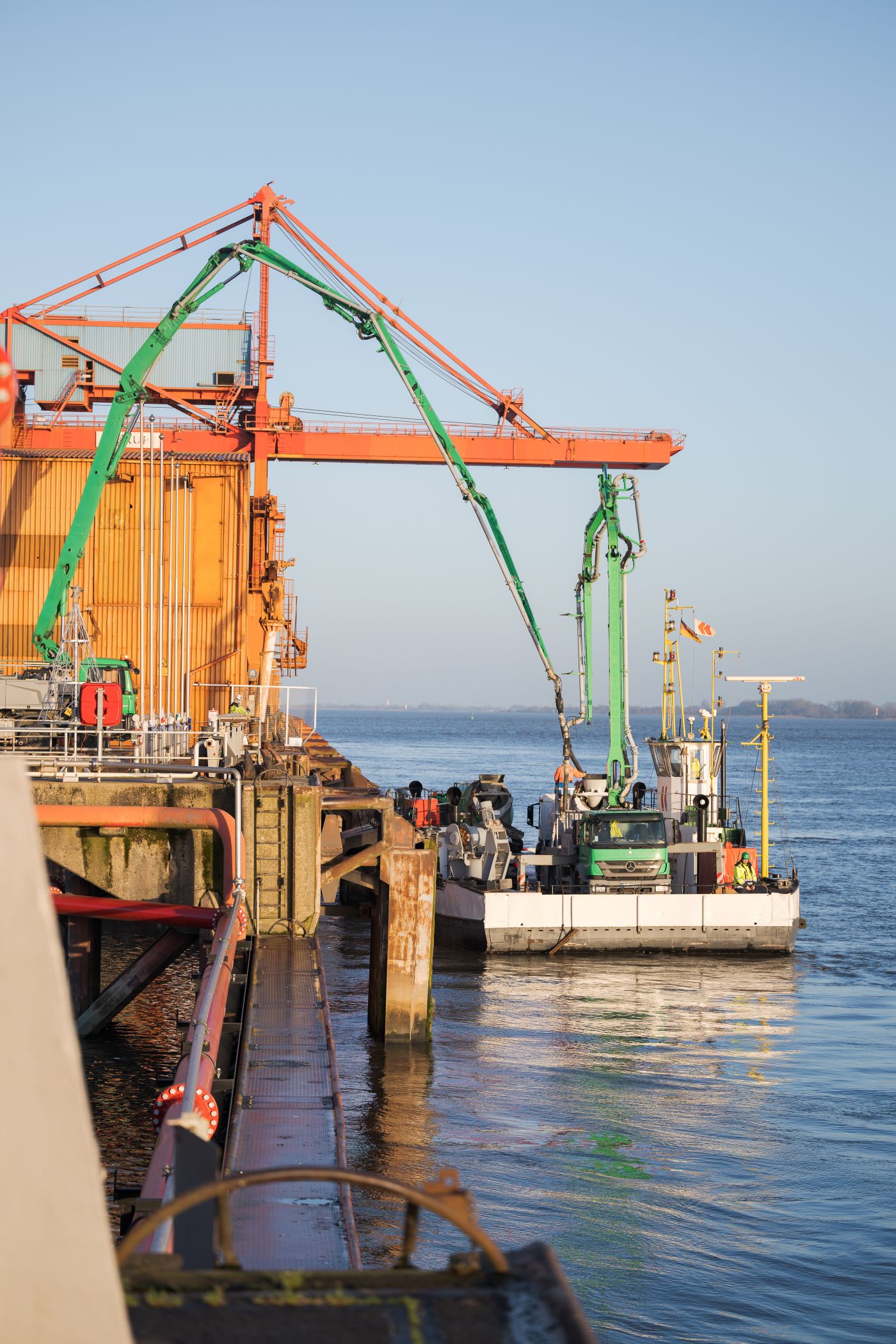 Industrieanwendung auf einem Gewässer: Eine Plattform mit gelben und orangefarbenen Strukturen steht links, während ein Schiff rechts anlegt. Eine grüne Betonpumpe verbindet beide. Klare Wetterbedingungen und ruhige Wasseroberfläche.