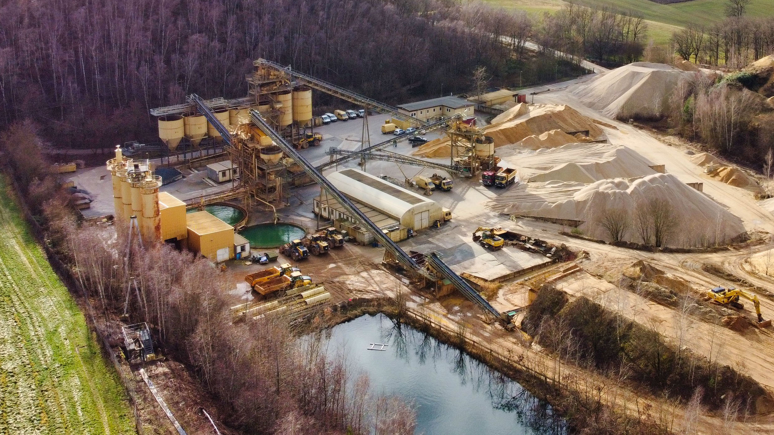 Luftaufnahme eines Kieswerks in ländlicher Umgebung, umgeben von Bäumen und Feldern. Sichtbar sind Förderbänder, Industriegebäude, Kieshalden, Fahrzeuge und Wasserbecken im Vordergrund.