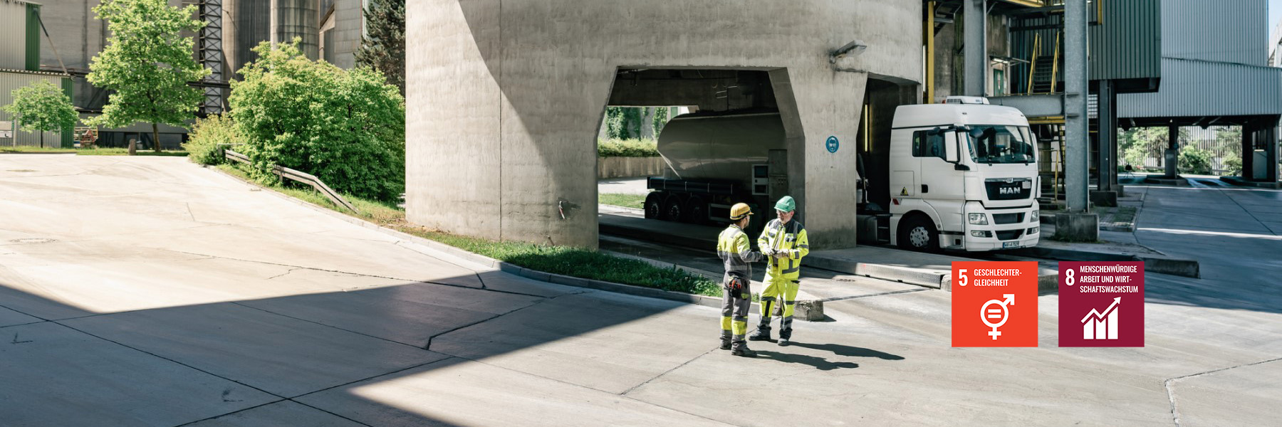 Zwei Arbeiter in Schutzkleidung stehen auf einem Zementwerk und unterhalten sich; im Hintergrund ein weißer Silozug.