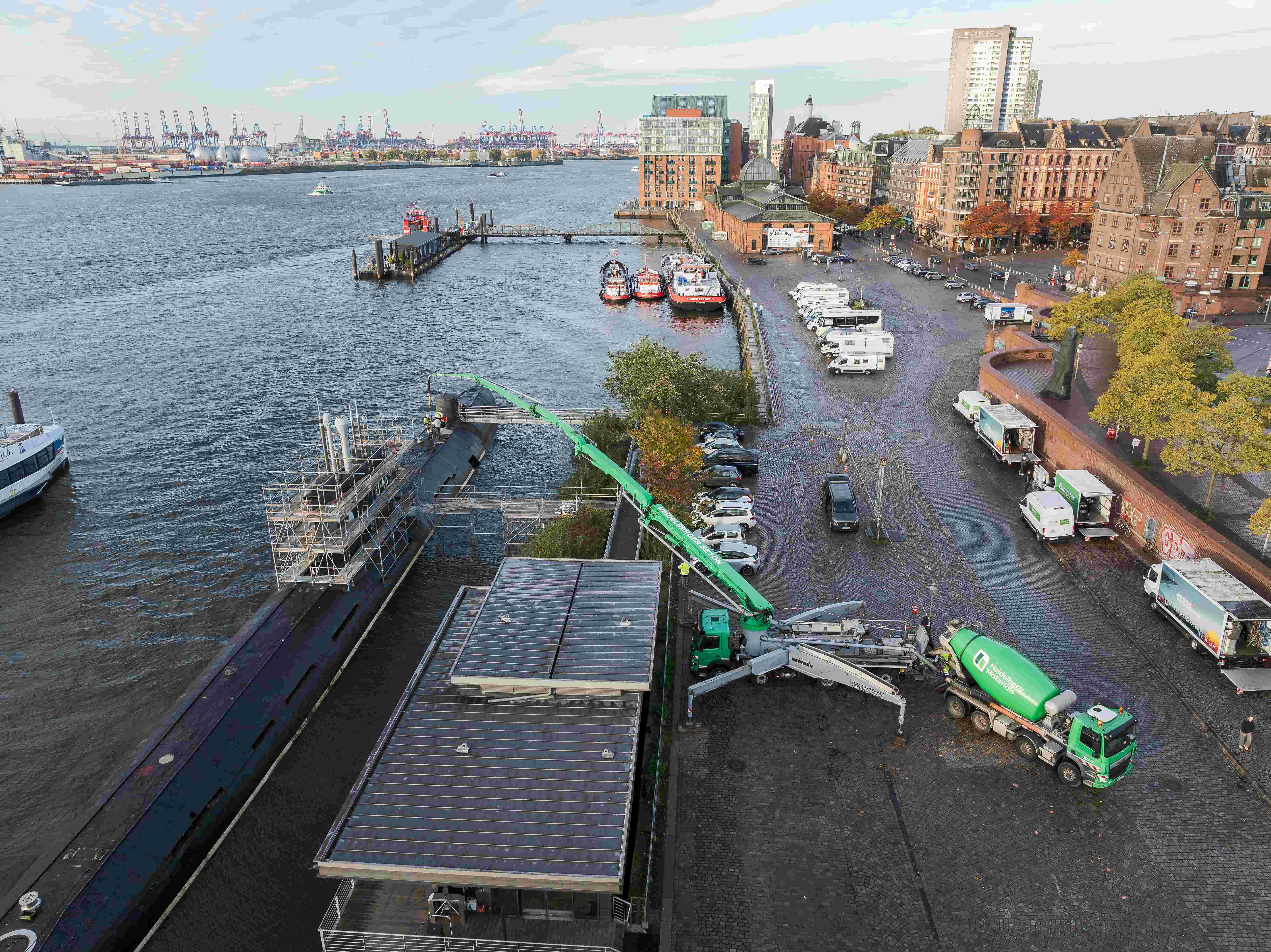 Eine Betonpumpe pumpt Beton in ein U-Boot am Ufer eines Flusses. Links der Hafen mit Booten, rechts eine Straße mit parkenden Fahrzeugen und Gebäuden im Hintergrund. Im Vordergrund eine grüne Betonpumpe, die in das Schiff ragt.