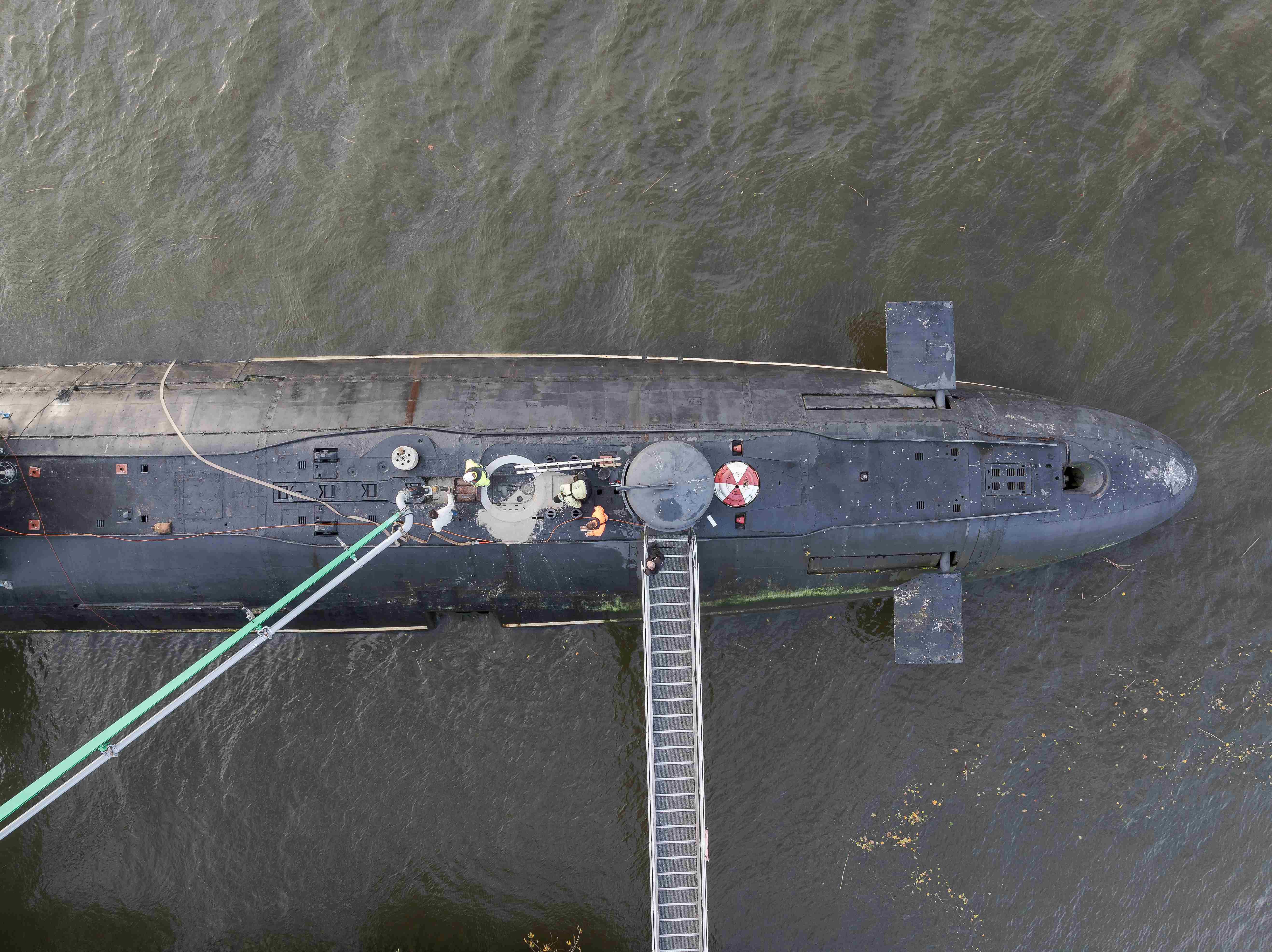 Luftaufnahme eines U-Boots, das seitlich an einem Steg im Wasser liegt. Zwei grüne Schläuche verlaufen von der Plattform zum U-Boot, dessen Oberdeck sowie ein geöffneter Einstieg sichtbar sind. Vier Personen stehen um den Einstieg herum. Es wird betoniert.