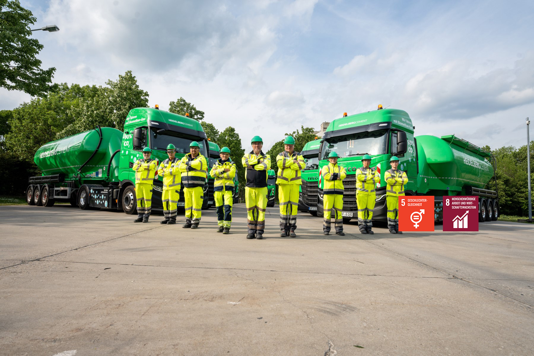 Gruppe von neun Personen in gelber Arbeitskleidung und Schutzhelmen vor zwei grünen Silozügen. Im Hintergrund sind Bäume und ein bewölkter Himmel sichtbar.