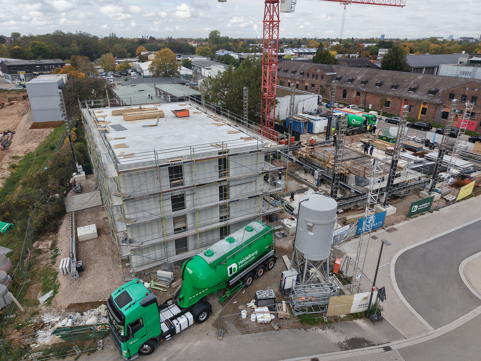 Luftaufnahme der Baustelle DREIHAUS mit einem teilweise errichteten mehrstöckigen Gebäude links, Baugerüsten rechts und einem Silozug von Heidelberg Materials im Vordergrund. Im Hintergrund sind weitere Gebäude, ein Baukran und Bäume sichtbar.
