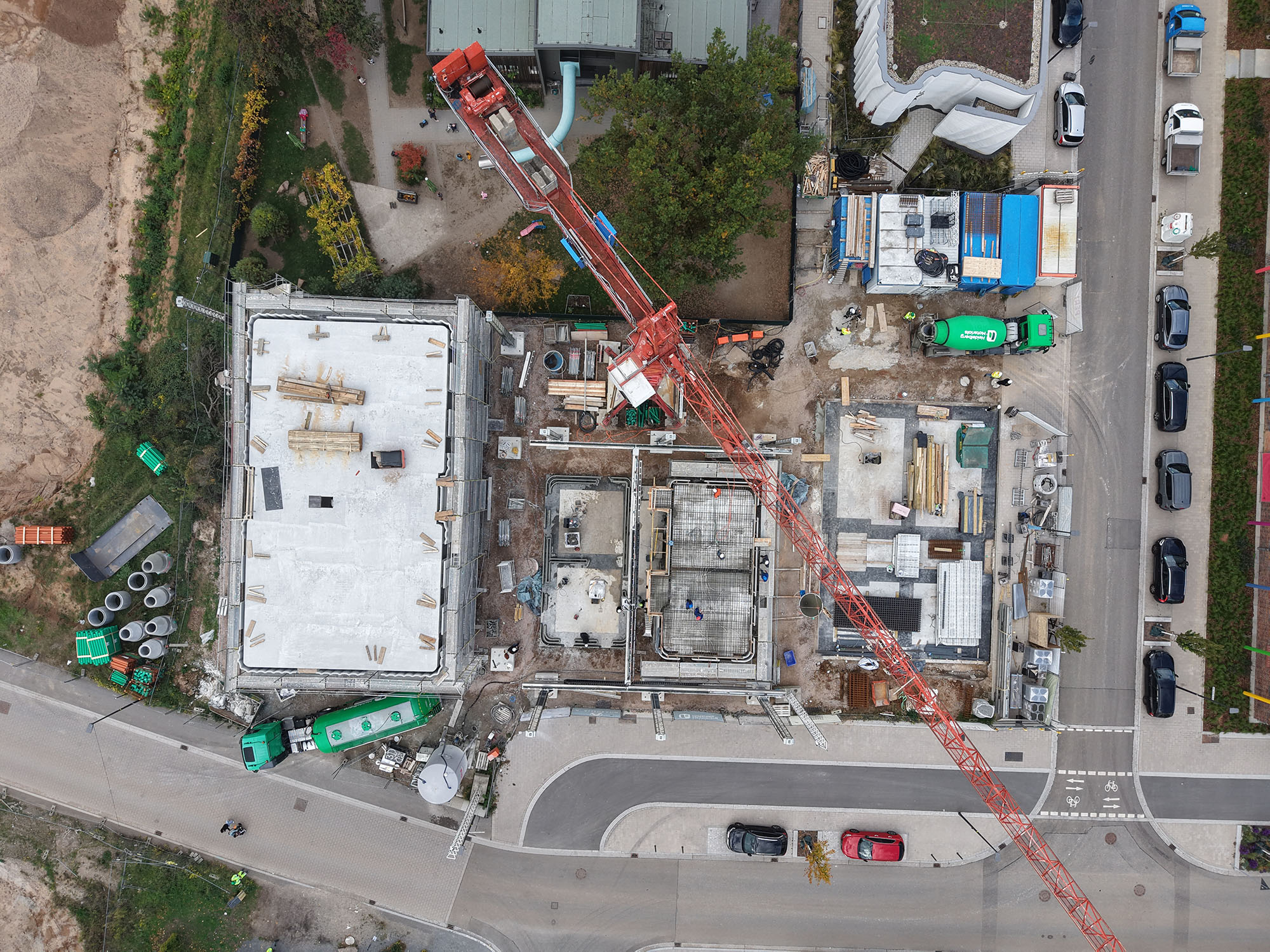 Baustelle aus der Vogelperspektive: Zwei Gebäude in unterschiedlichen Bauphasen, ein roter Baukran überspannt die Fläche. Links ein fast fertiges Gebäude mit weißem Dach, rechts sind Fundamente und Rohbauarbeiten sichtbar. Umgeben von Straßen mit geparkten Autos und Baucontainern.