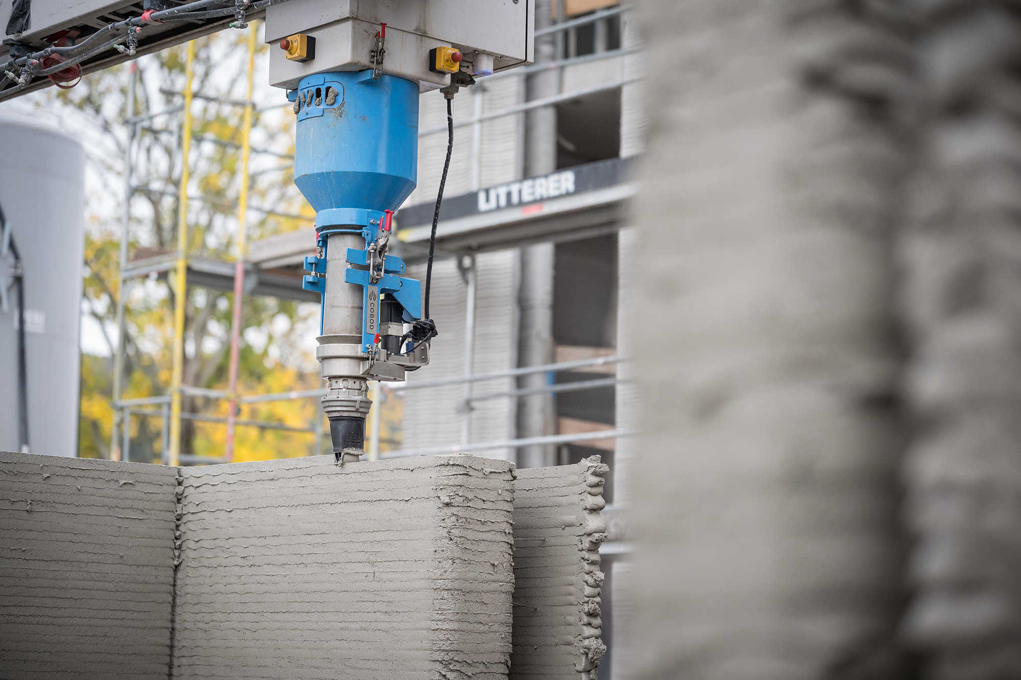 3D-Drucker beim Bau eines Gebäudes: Eine blaue Druckerkomponente extrudiert grauen Beton in Schichten, um eine Wandstruktur aufzubauen. Im Hintergrund sind Gerüste und ein Baum sichtbar.