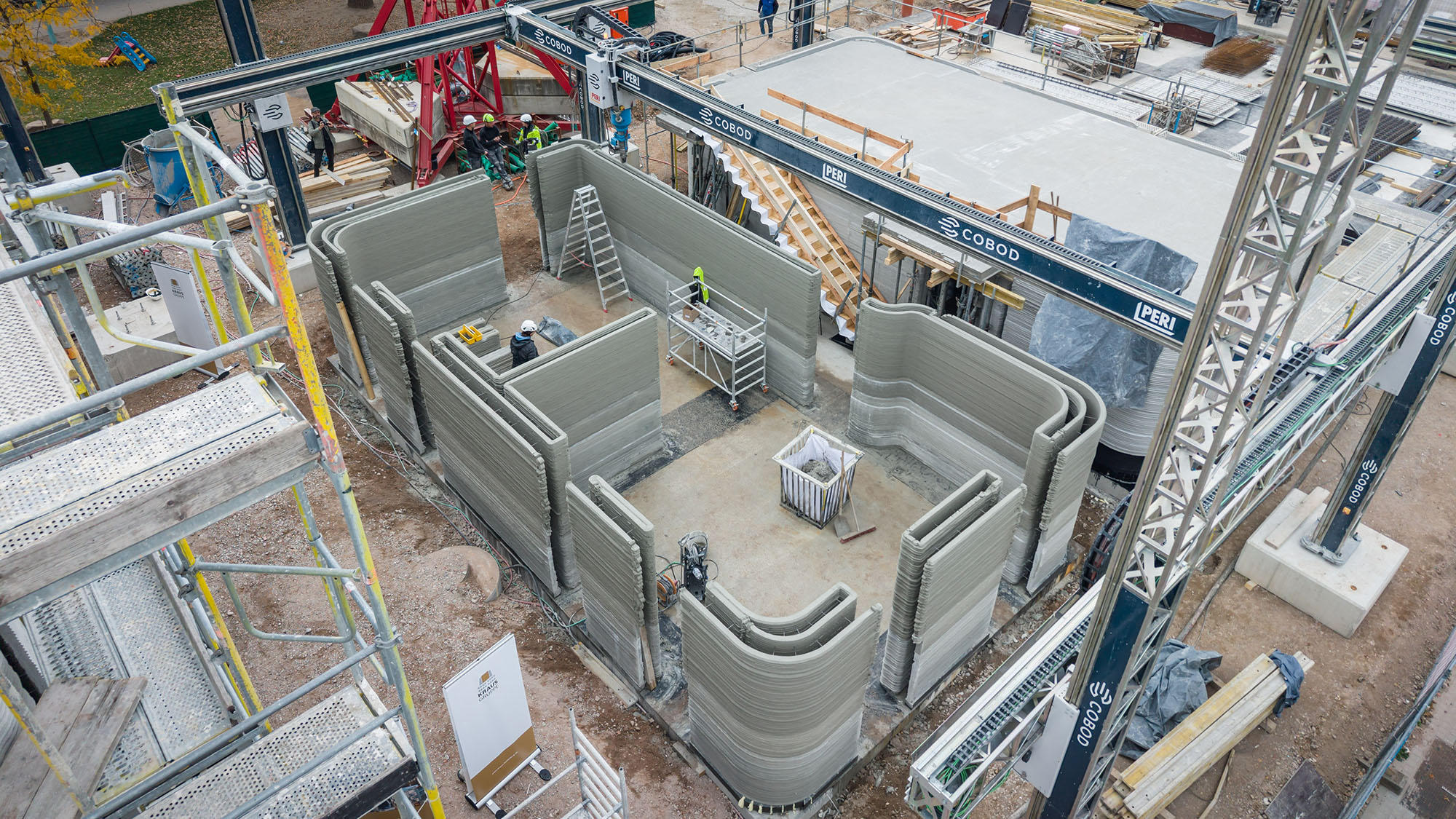 Luftaufnahme einer Baustelle, auf der ein Haus mit einem 3D-Drucker gebaut wird. Gedruckte Wände in grauer Farbe formen bereits Räume. Arbeiter in Schutzkleidung stehen im Inneren und am Rand, während Baugeräte und Gerüste das Gelände umgeben. Oben rechts sind Baustellenelemente wie Betonplatten sichtbar.