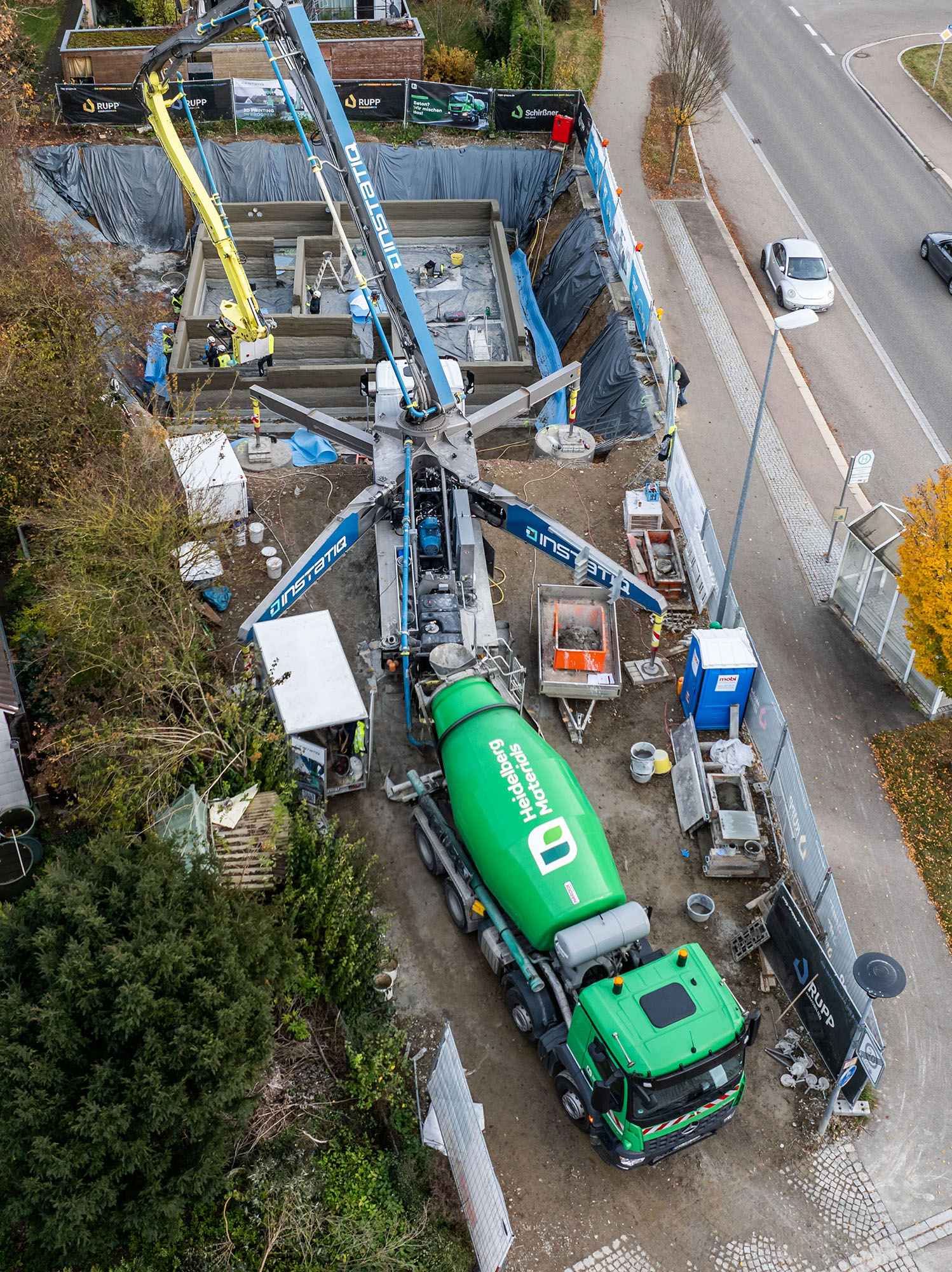 Baustelle aus der Vogelperspektive. Im Zentrum ein großer Betonförderarm, der über die Baugrube ragt. Unten ein grüner Betonmischer, der Beton liefert. Arbeiter und Baugeräte sind umgeben von Absperrzäunen. Rechts oben eine Straße mit vorbeifahrenden Autos.