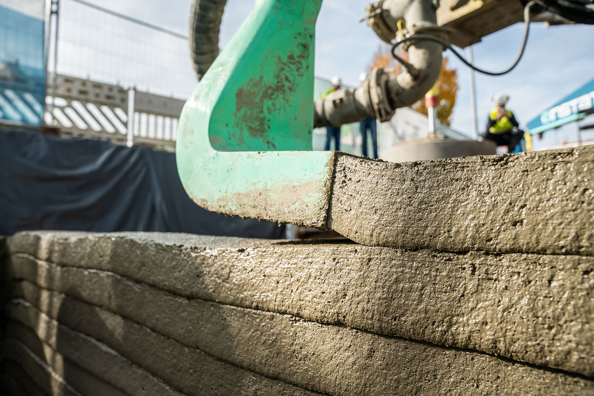 Nahaufnahme eines 3D-Druckers, der Schichten aus Beton aufeinanderlegt. Im Hintergrund sind Baugerüste, Absperrungen und Arbeiter mit Schutzwesten sichtbar.