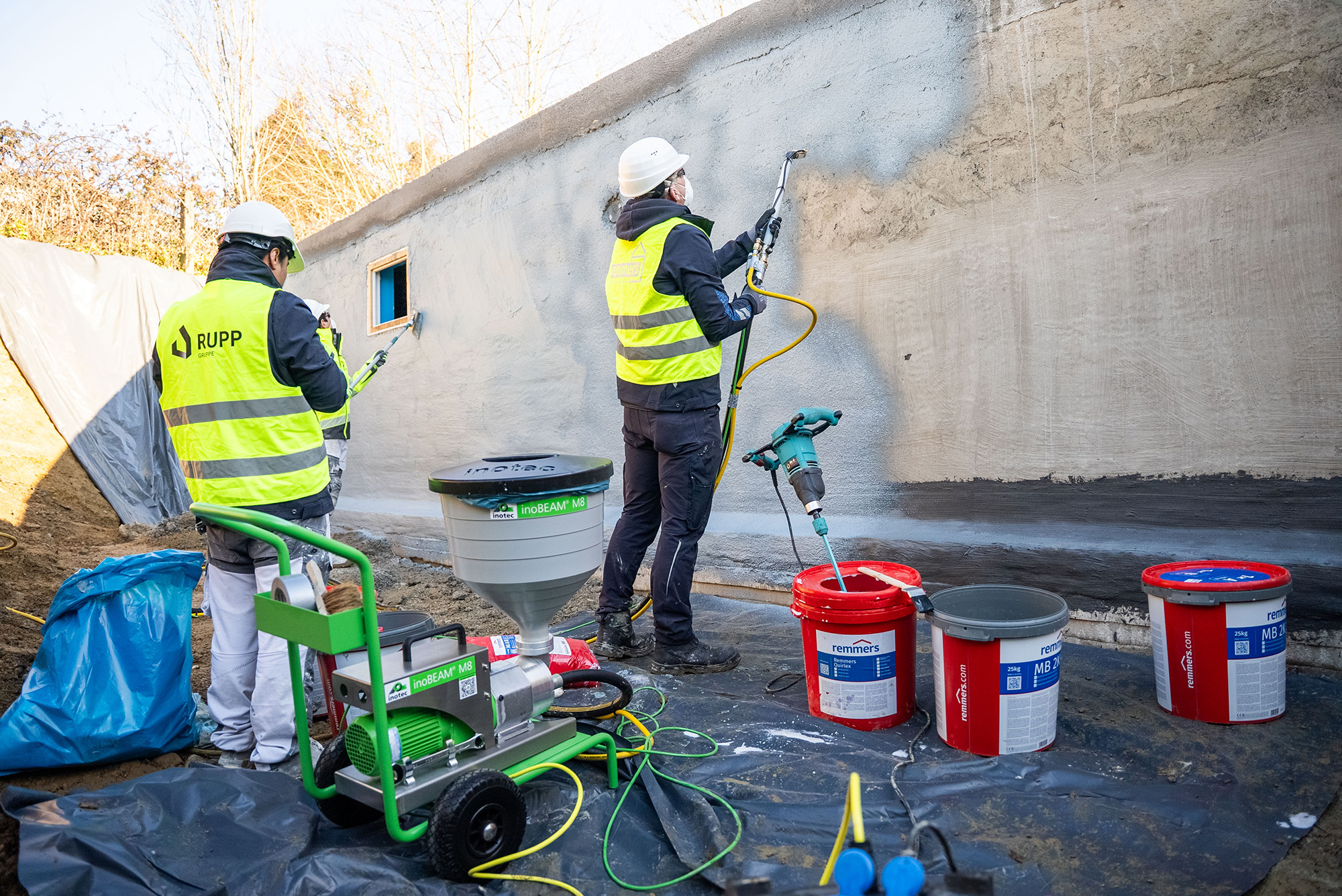Zwei Arbeiter in gelben Warnwesten und weißen Helmen sorgen für die Bauwerksabdichtung. Die Umgebung zeigt einen Baustellenbereich im Freien.