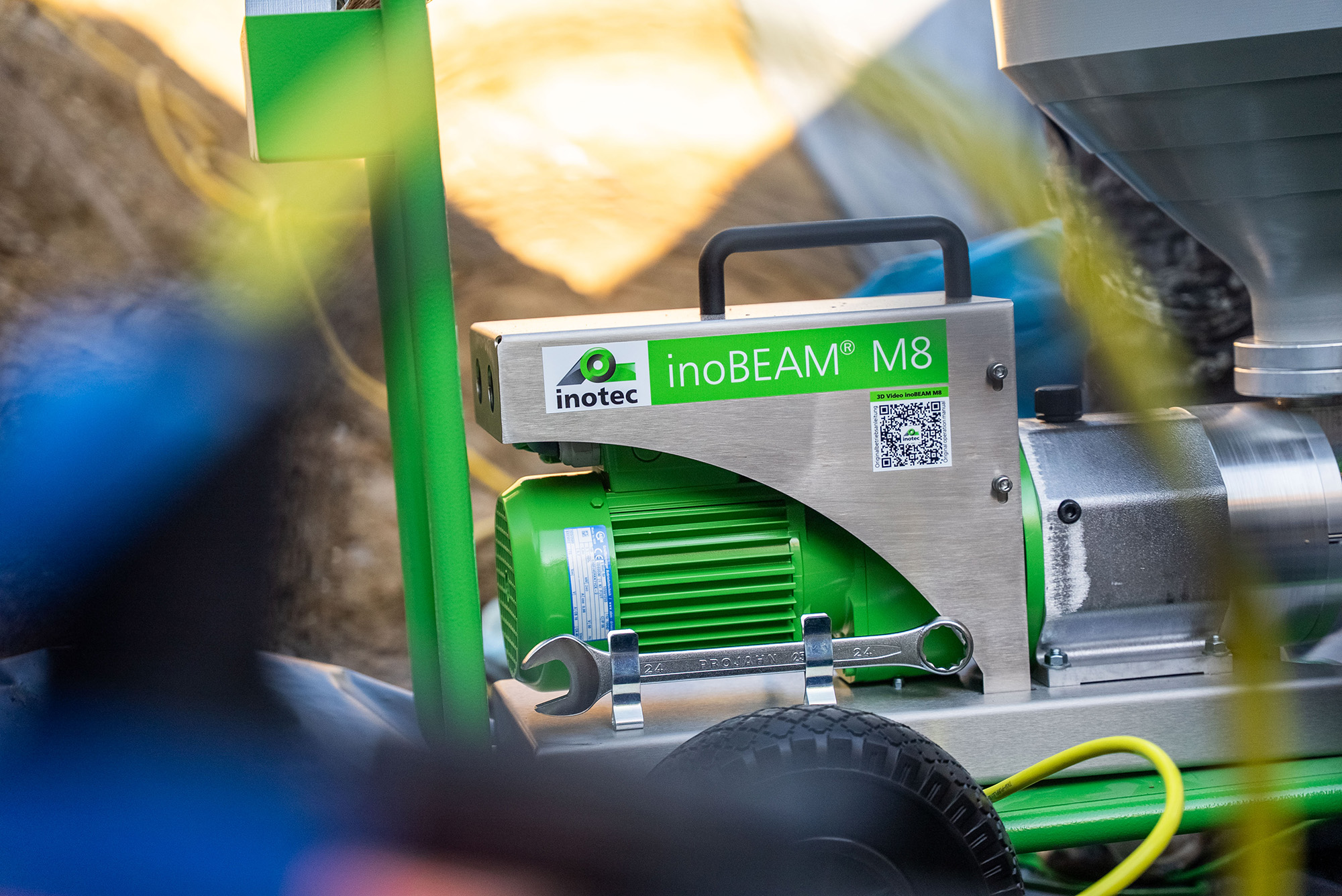 Grüne InoBEAM® M8-Maschine mit seitlichem QR-Code und sichtbarem Logo des Herstellers. Im Hintergrund verschwommenes Material und Schläuche, die auf eine Baustellenumgebung hinweisen.