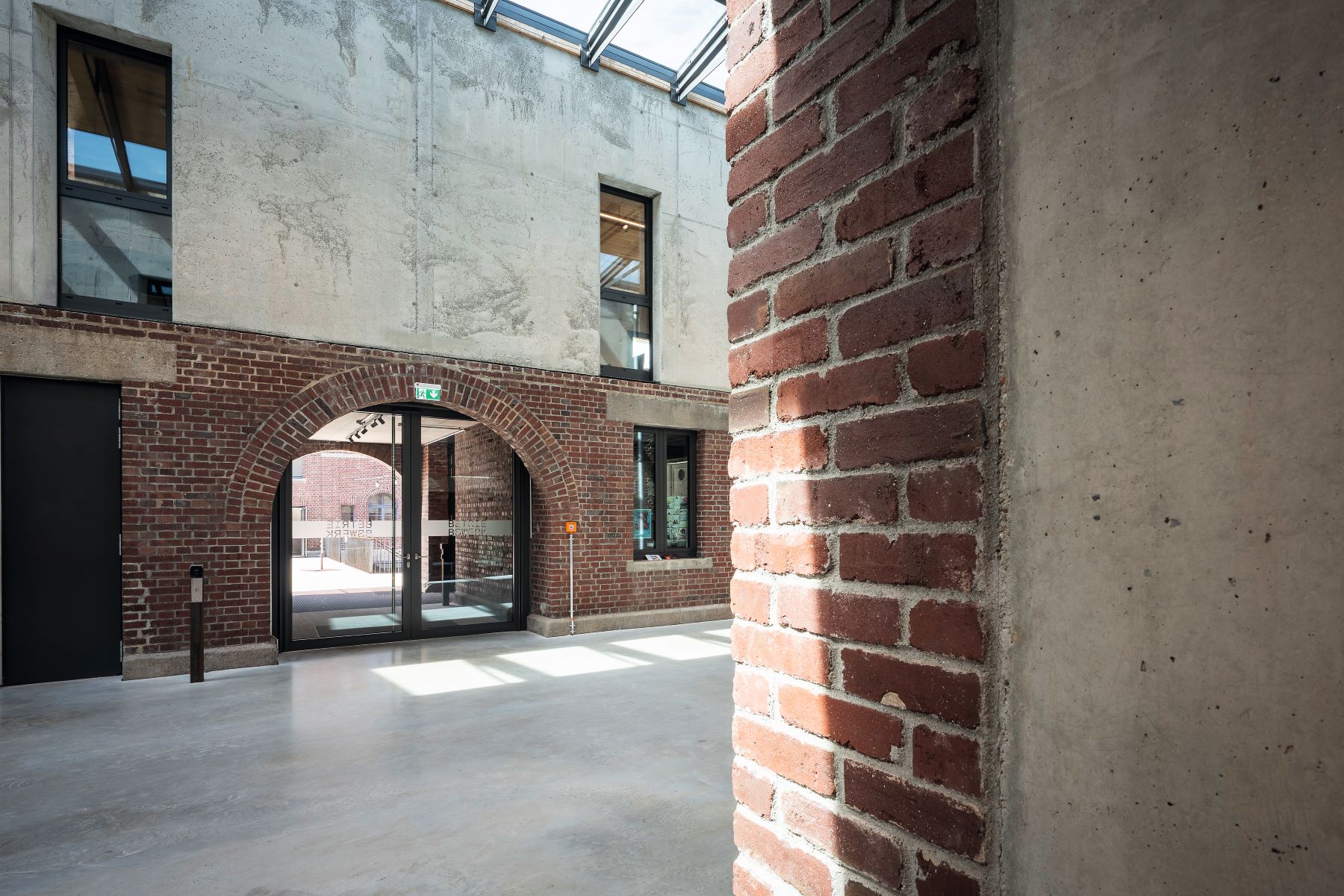 Innenraum mit Backsteinmauer und Sichtbeton; natürlichem Licht fällt durch ein Glasdach. Ein gewölbter Backsteindurchgang führt zu großen Glastüren, die nach draußen führen.