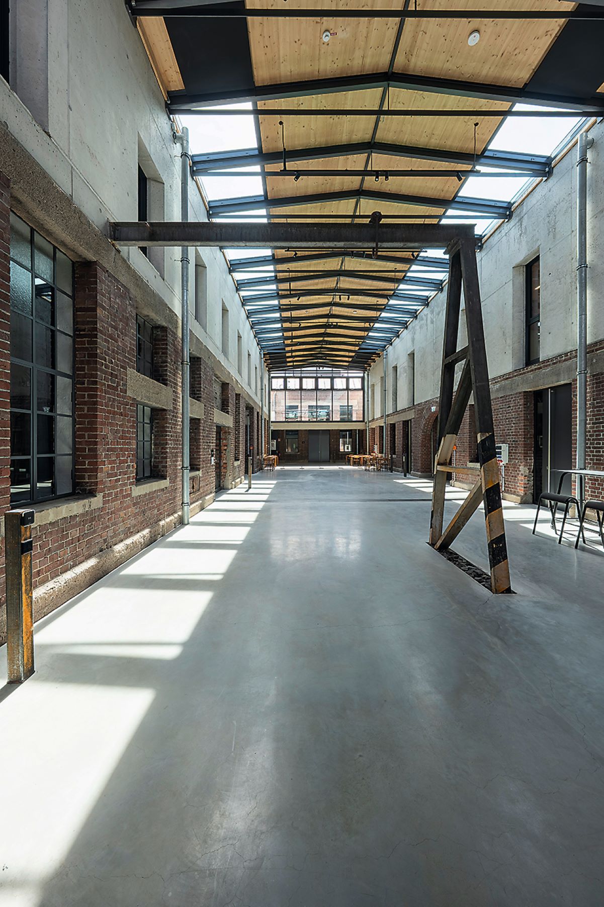 Lange Halle mit glasgedecktem Dach, freiliegendem Backsteinmauerwerk und Sichtbetonflächen. Tageslicht fällt durch große Glasflächen auf den glatten Boden. Links und rechts befinden sich Fenster- und Türöffnungen; im Hintergrund stehen Tische und Stühle. Ein dunkles Stahlgestellt steht im vorderen Bereich der Halle.